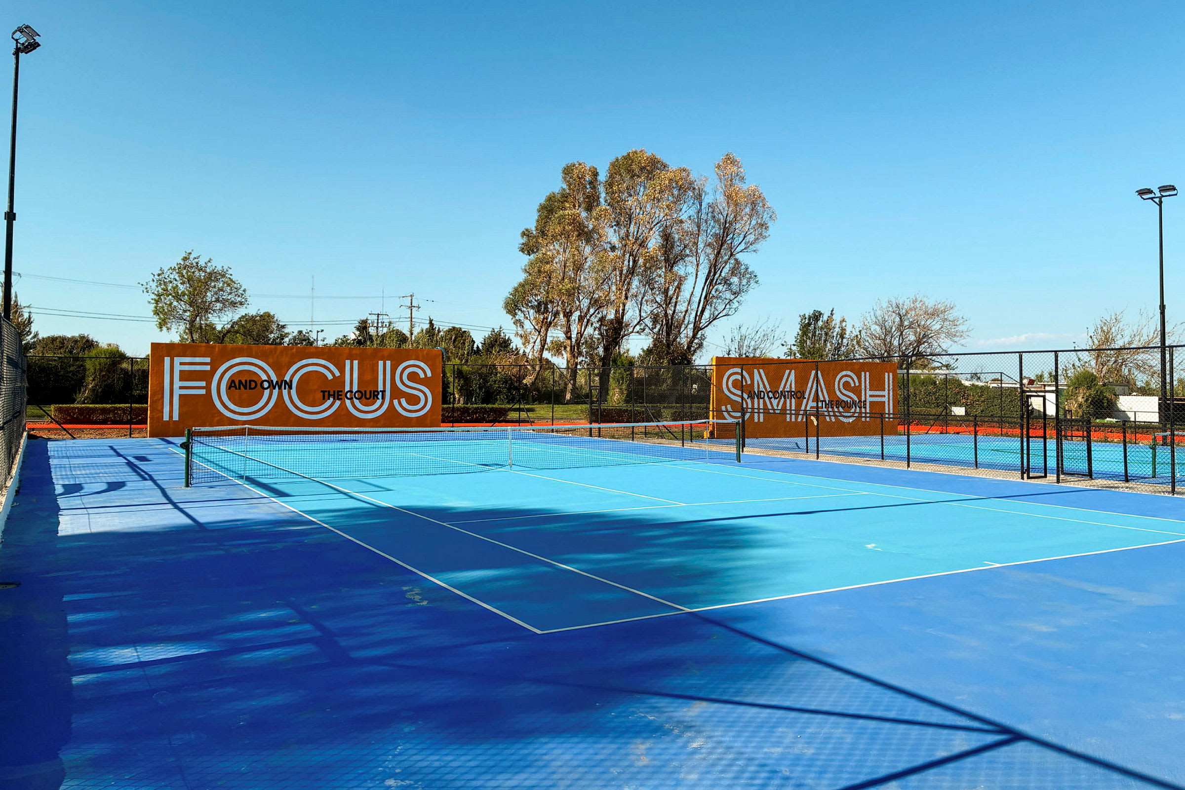 Sininen tenniskenttä ulkona, taustalla puita ja kylttejä, joissa lukee "Focus" ja "Smash". (Tekoälyn luoma teksti)