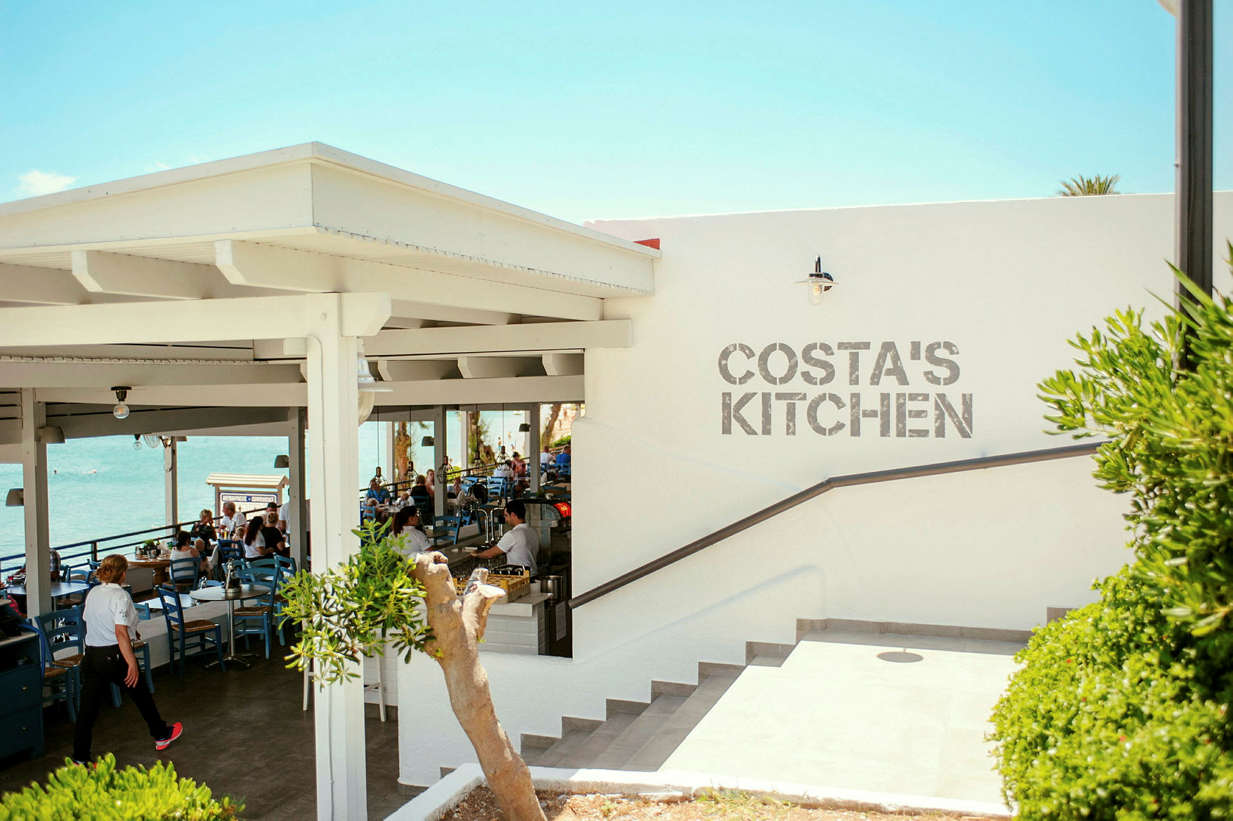 Ulkoterassiravintola, jossa on merinäköala ja seinässä teksti 'Costa's Kitchen'. (Tekoälyn luoma teksti)