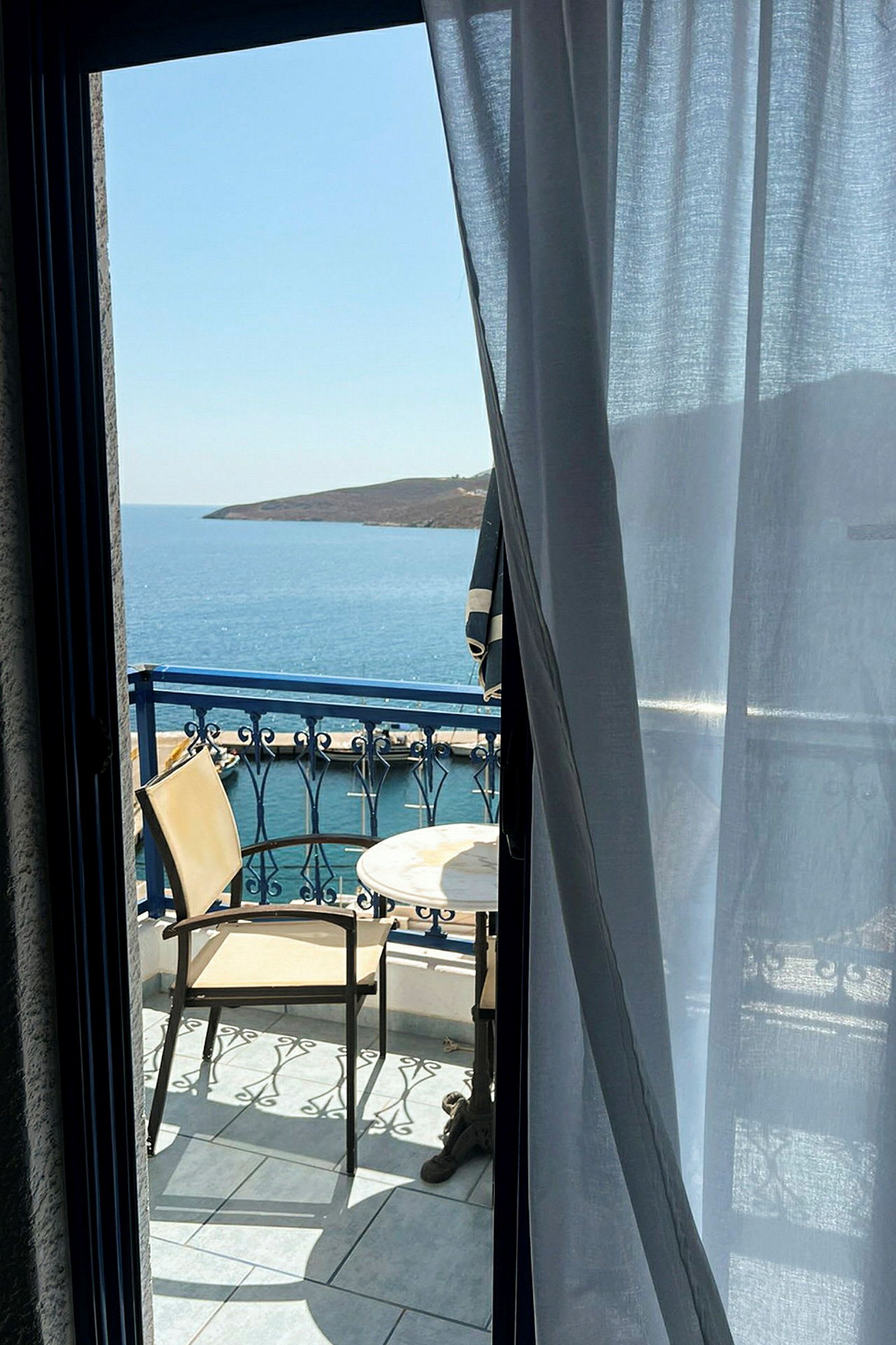 Näkymä huoneesta parvekkeelle, jossa kaksi tuolia ja pöytä, taustalla meri ja rannikko. (Tekoälyn luoma teksti)