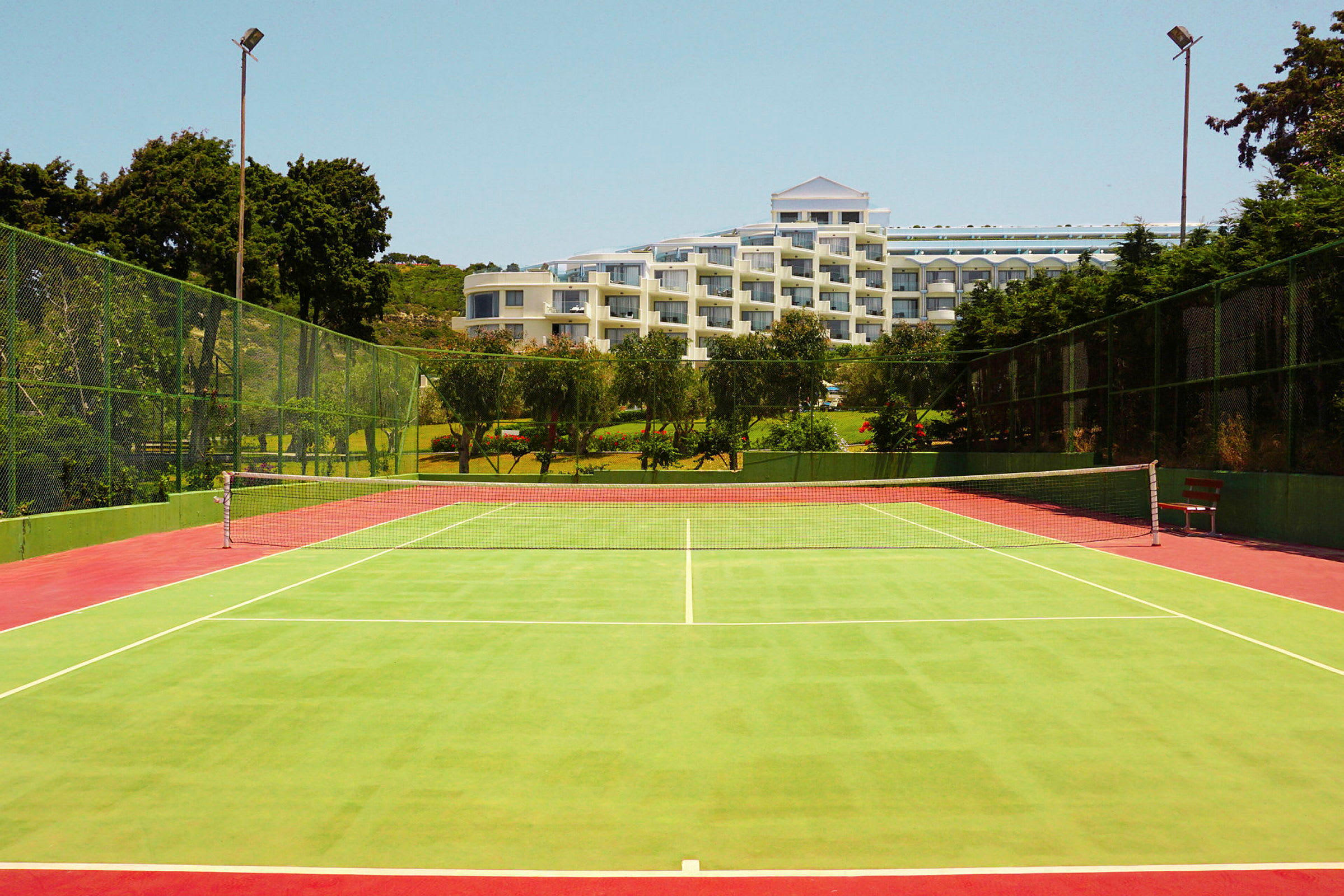 Vihreä tenniskenttä, jonka keskellä on verkko, ja taustalla on hotelli, jota ympäröivät puut. (Tekoälyn luoma teksti)