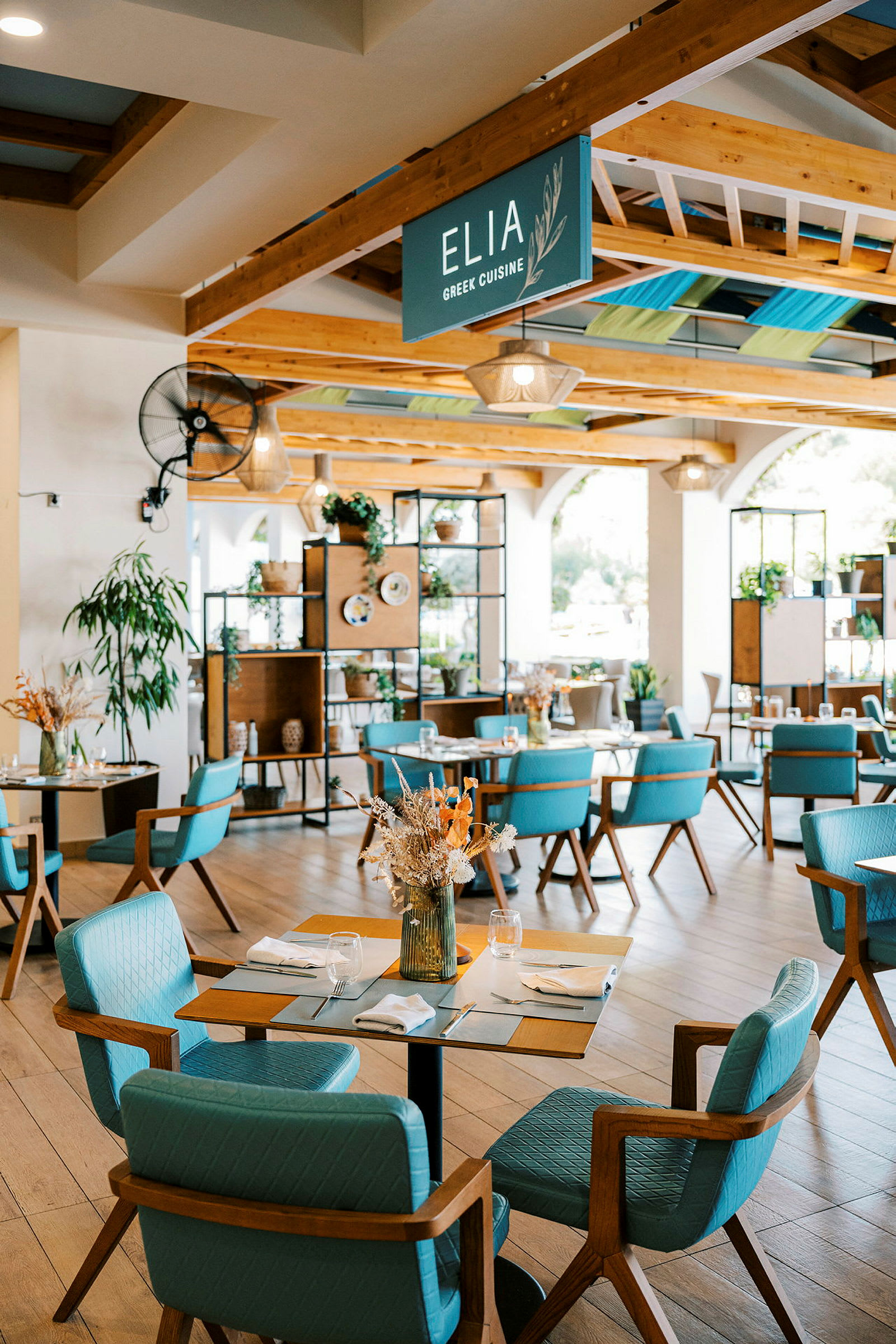 Valoisa ja moderni ravintola, jossa on puuyksityiskohtia ja siniset tuolit. Koristeltu kasveilla ja kuivatuilla kukka-asetelmilla. (Tekoälyn luoma teksti)