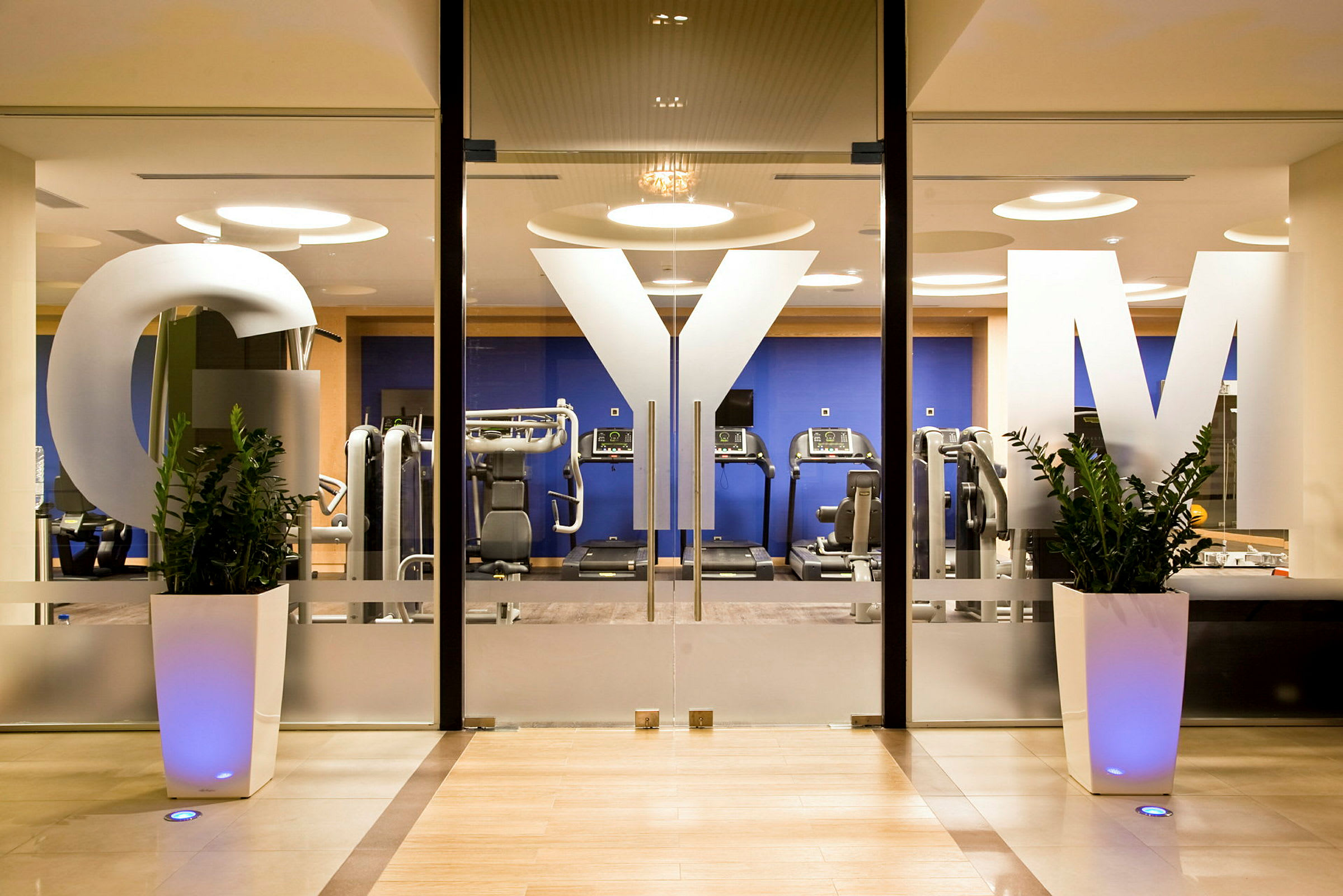 Kuntosalin sisäänkäynti lasiovilla, joissa lukee "GYM", ja taustalla näkyy laitteita ja harjoitusvälineitä. (Tekoälyn luoma teksti)