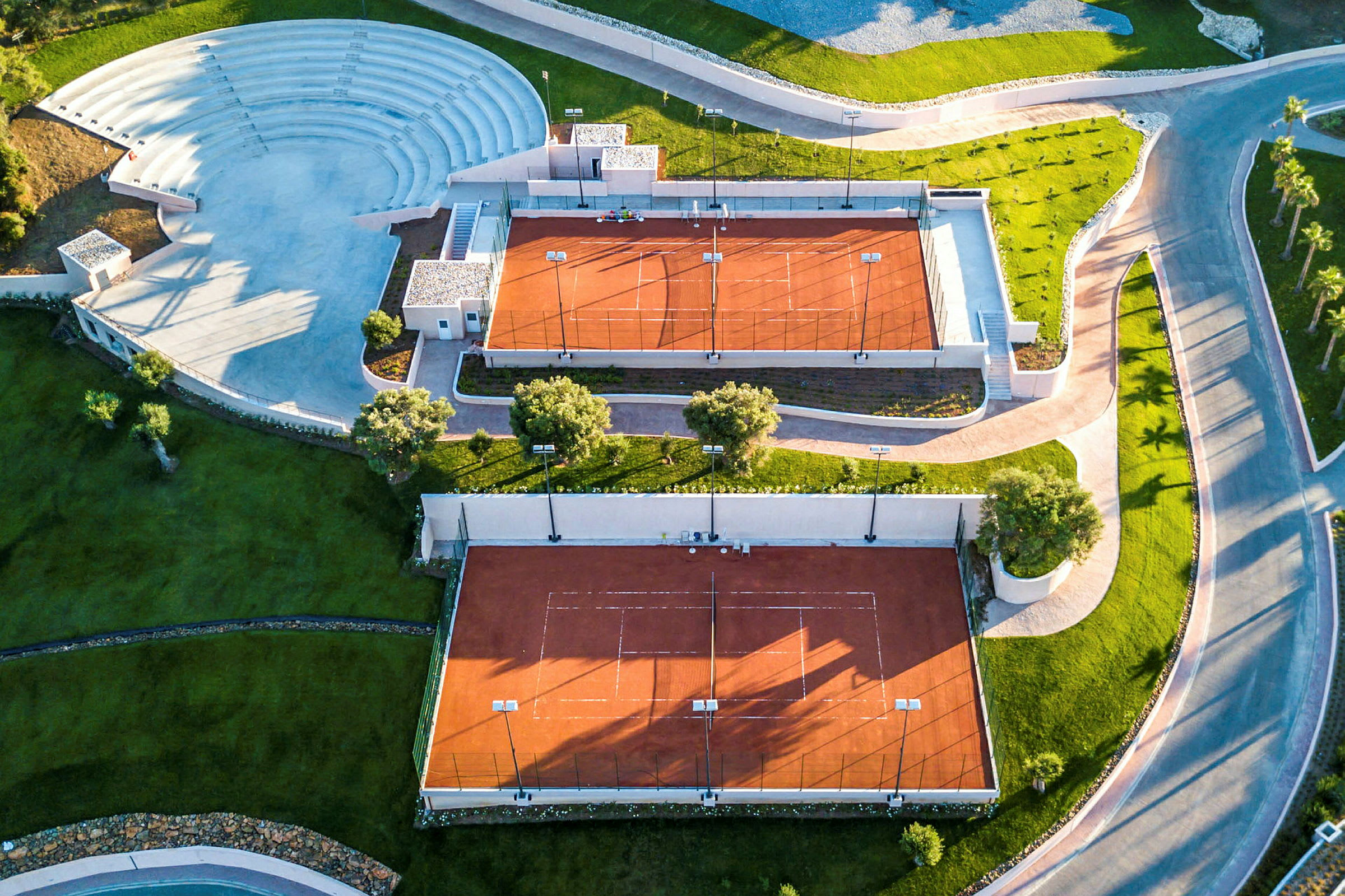 Ilmakuva kahdesta punaisella kentällä varustetusta tenniskentästä ja viereisestä ulkoilma-amfiteatterista. (Tekoälyn luoma teksti)