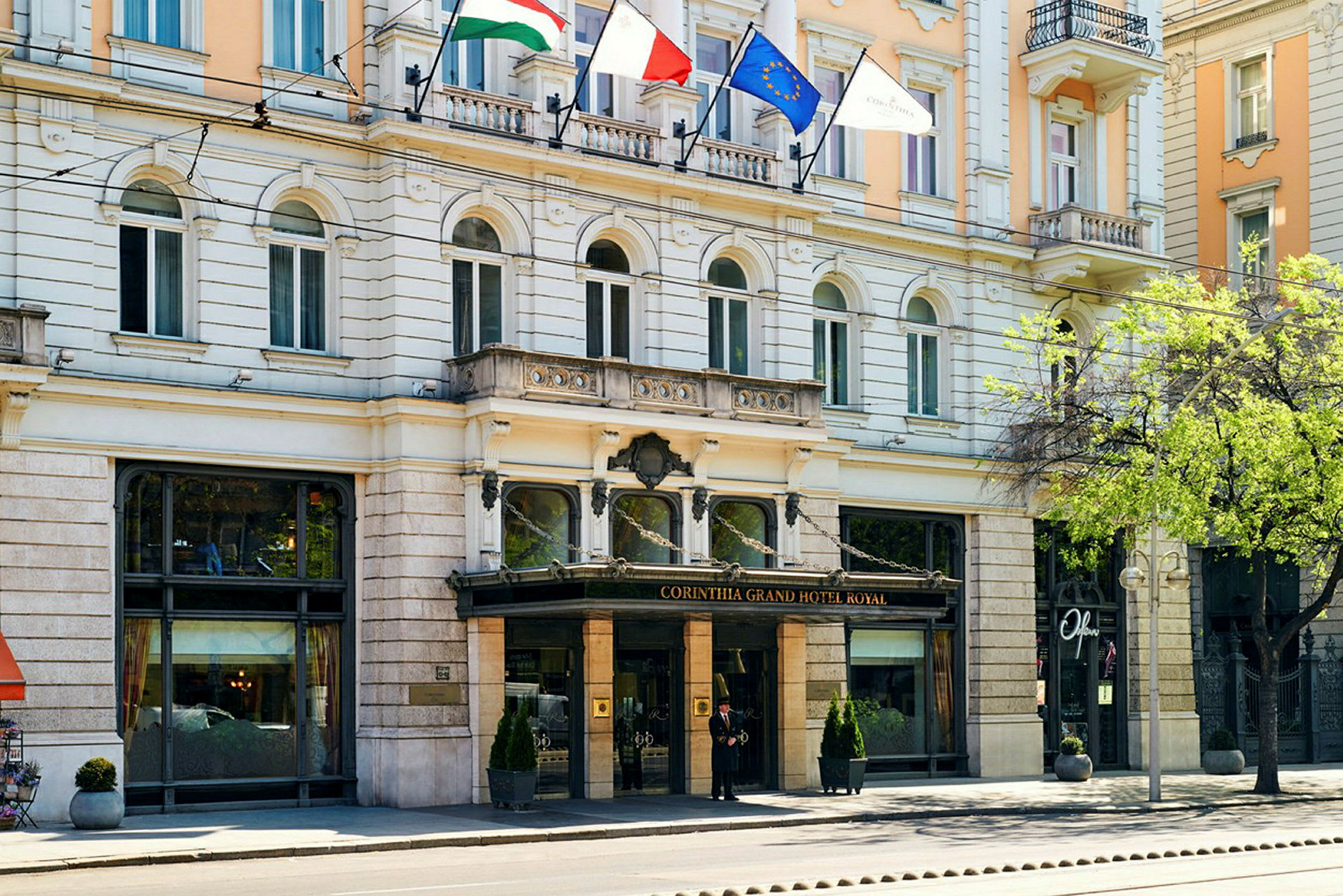 Corinthia Grand Hotel Royal -hotellin etupuoli, jossa lasisisäänkäynti ja liput yllä. (Tekoälyn luoma teksti)