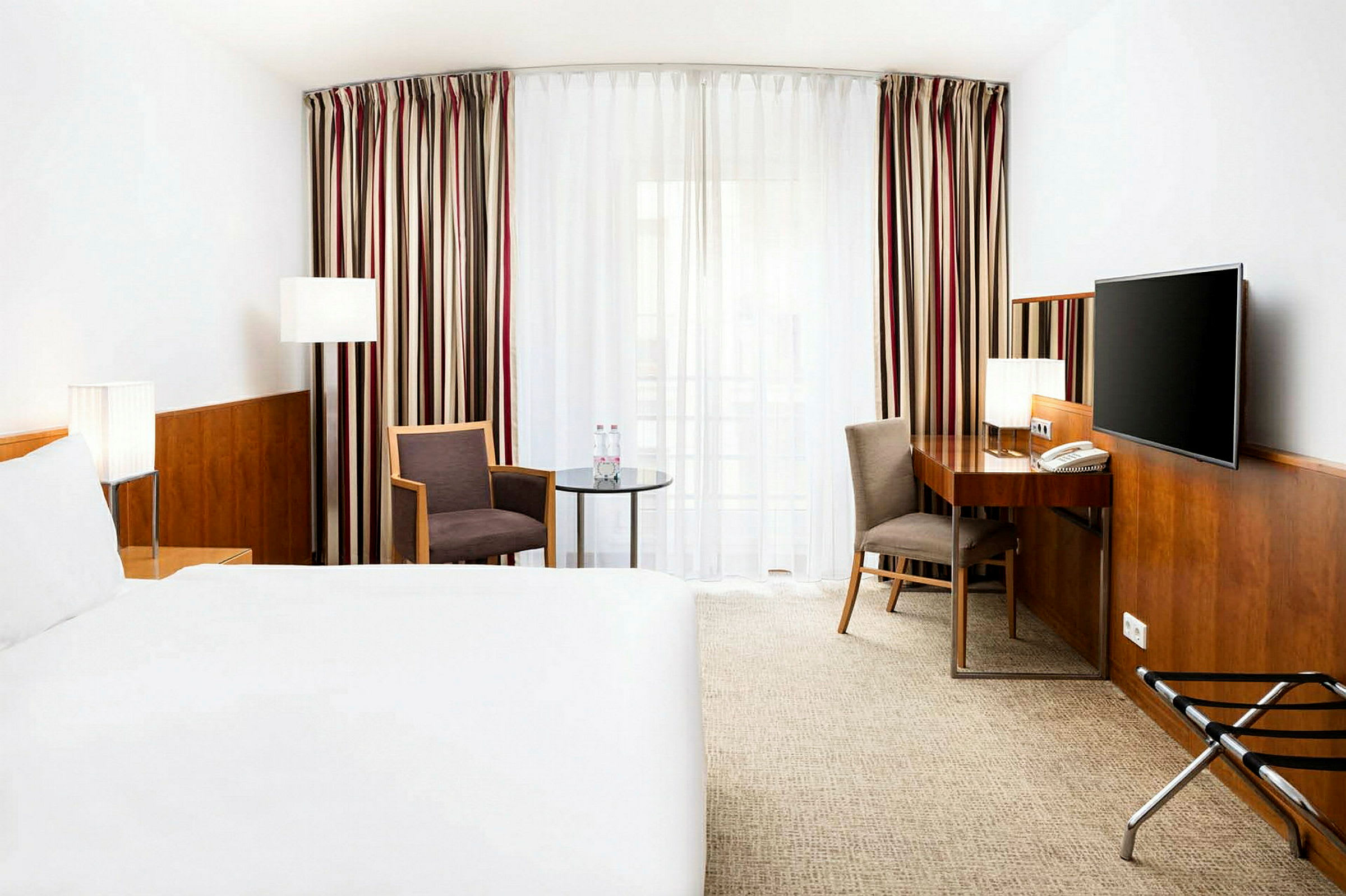 Hotellihuone, jossa on suuri sänky, kirjoituspöytä, kaksi tuolia ja televisio. Ikkuna on peitetty raidallisilla verhoilla. (Tekoälyn luoma teksti)