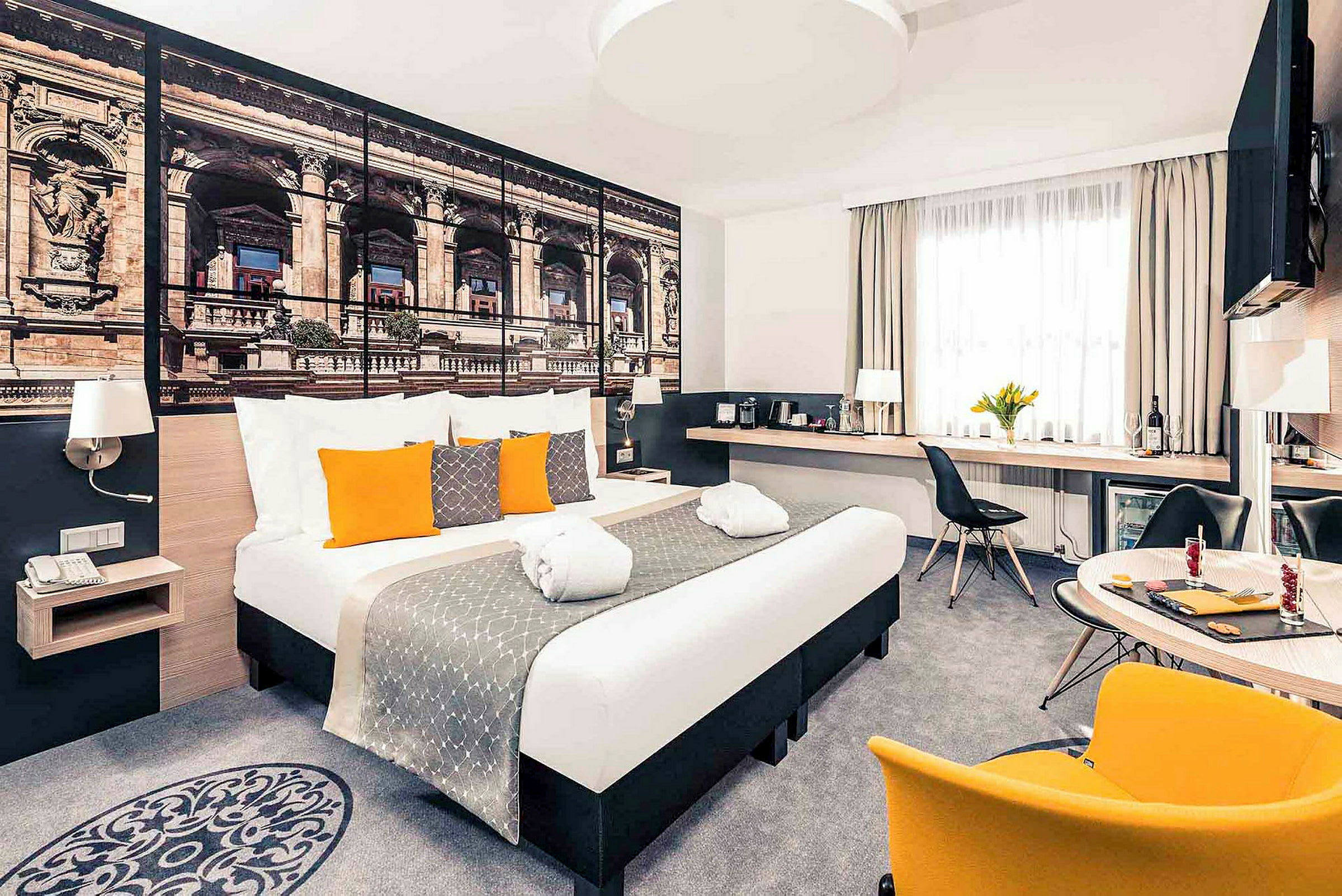 Moderni hotellihuone, jossa parisänky, koristeellisia tyynyjä ja suuri seinäkuva arkkitehtuurista. (Tekoälyn luoma teksti)