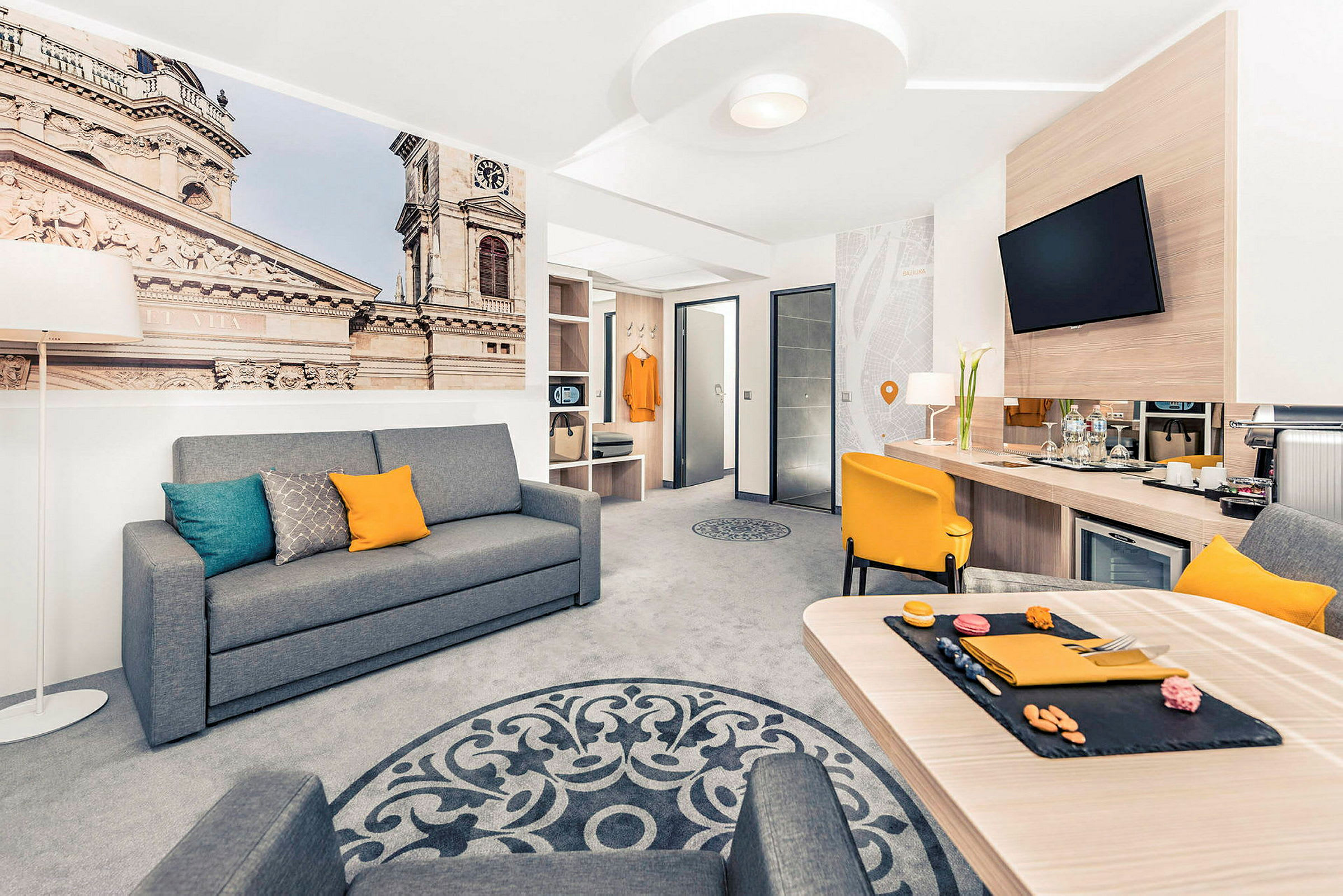 Modernin hotellihuoneen sisustus harmaalla sohvalla, keltaisella tuolilla ja pöydällä sekä suuri seinäkuva, joka esittää rakennusta. (Tekoälyn luoma teksti)