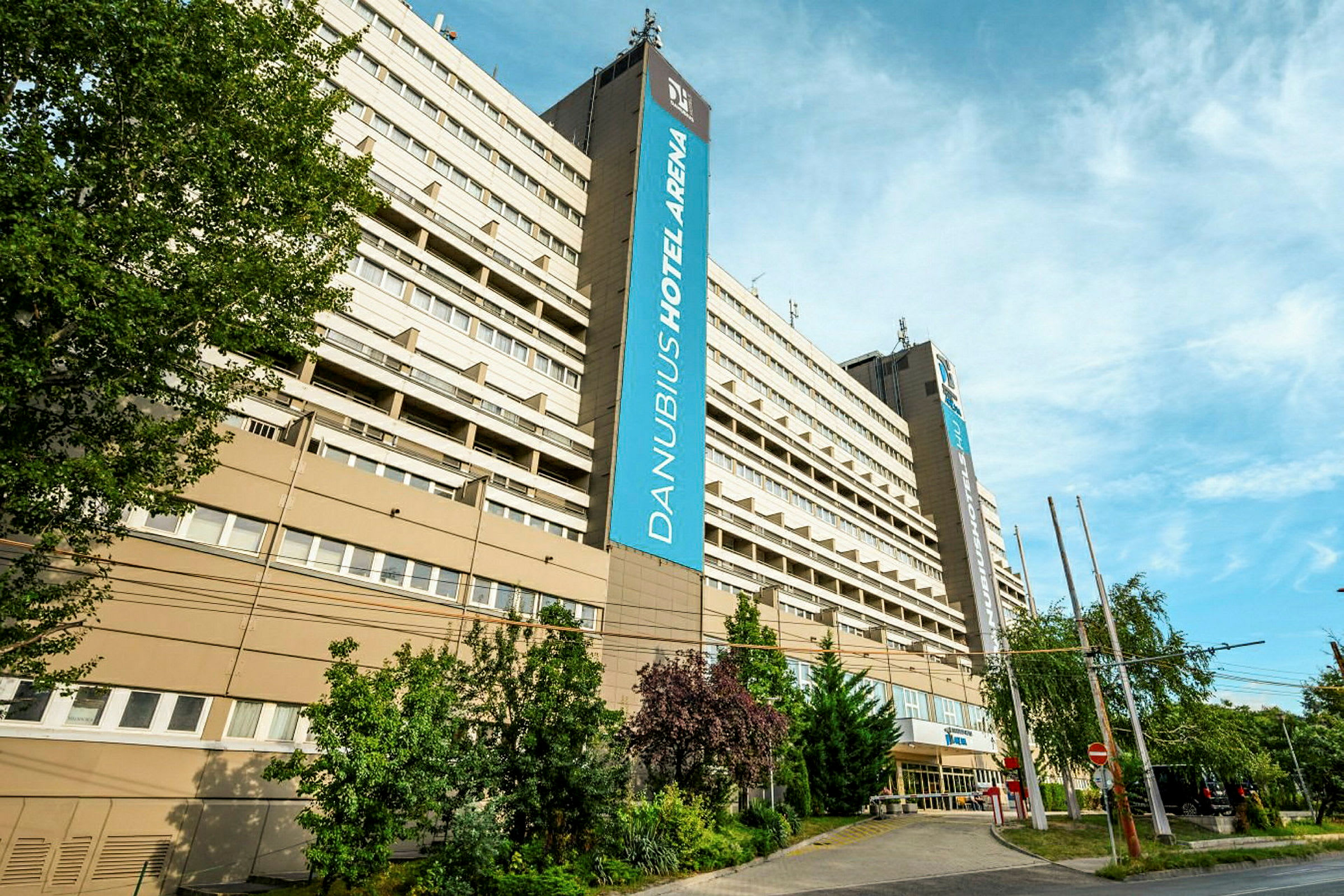 Suuri rakennus, jossa näkyy hotellin nimi 'Danubius Hotel Arena' kyltissä. Puita ja kasvillisuutta ympäröi alue, ja yllä näkyy sininen taivas. (Tekoälyn luoma teksti)
