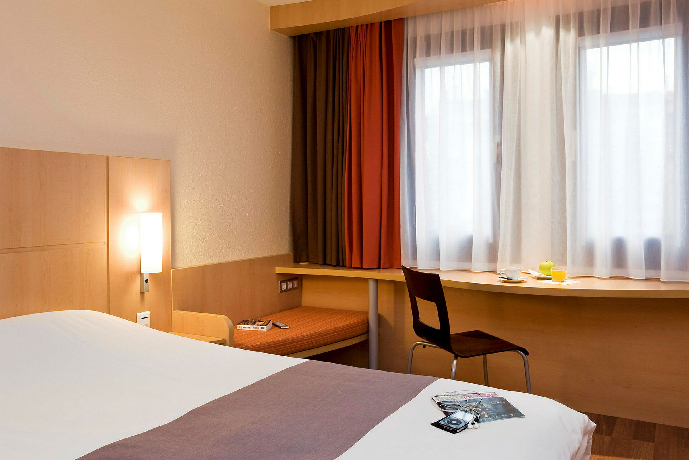 Hotellihuone, jossa on sänky, kirjoituspöytä ikkunan alla ja tuoli. Sänky on pedattu, ja kirjoituspöydällä on kuppi, lasillinen appelsiinimehua ja vihreä hedelmä. (Tekoälyn luoma teksti)