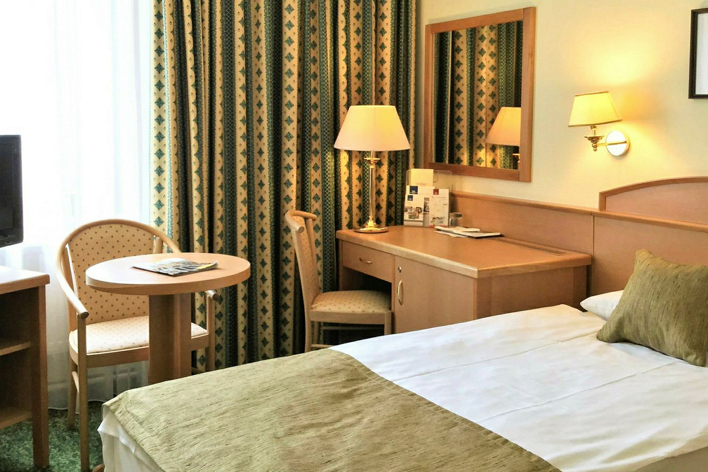 Hotellihuone, jossa on sänky, kirjoituspöytä ja istuin pienen pöydän ääressä. (Tekoälyn luoma teksti)