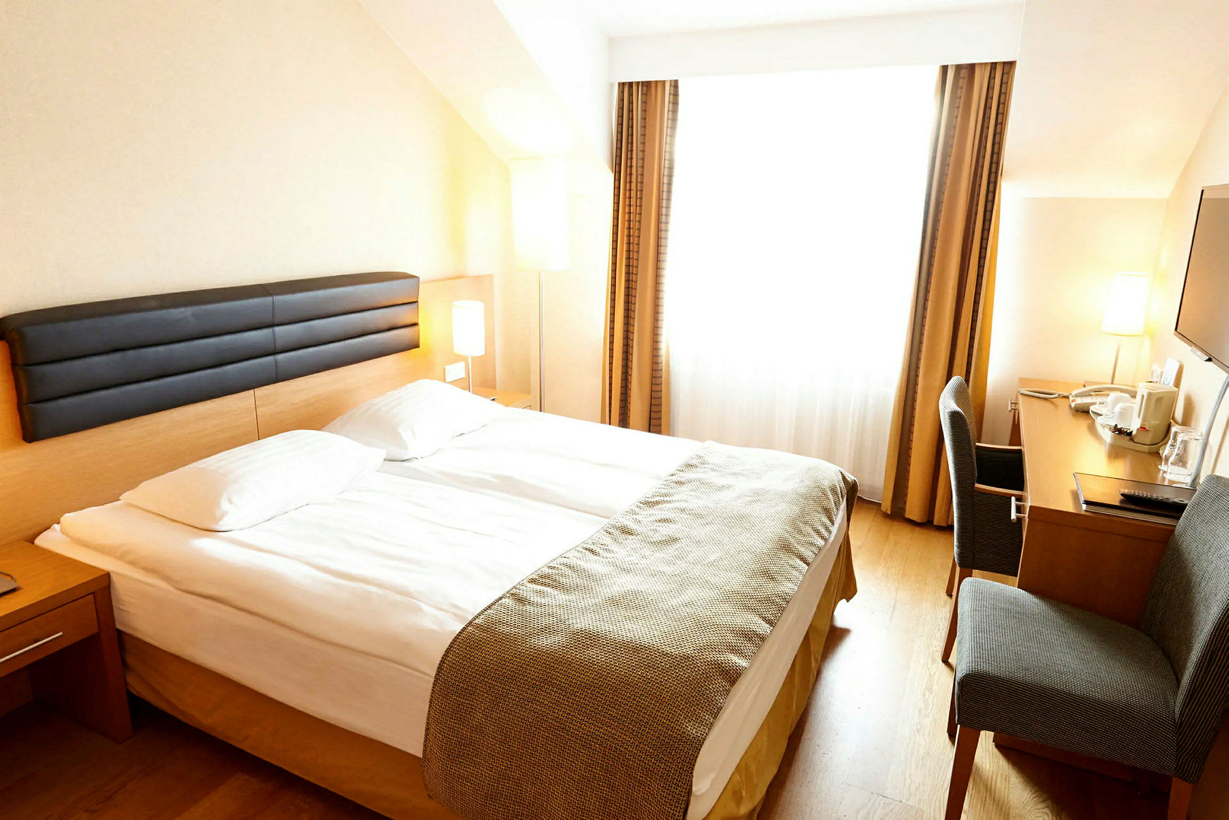 Hotellihuone, jossa on parivuode, kirjoituspöytä ja tuoli. Huoneessa on suuret ikkunat verhoineen. (Tekoälyn luoma teksti)