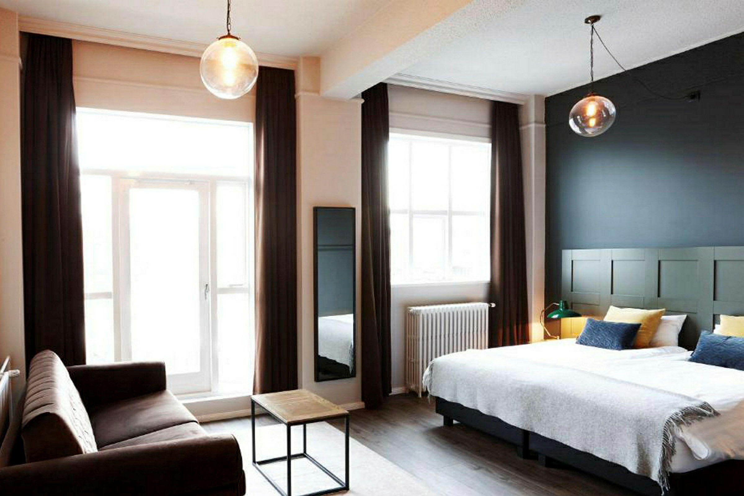 Hotellihuone, jossa on parivuode, sohva ja pieni sohvapöytä sekä isot ikkunat verhoilla. (Tekoälyn luoma teksti)