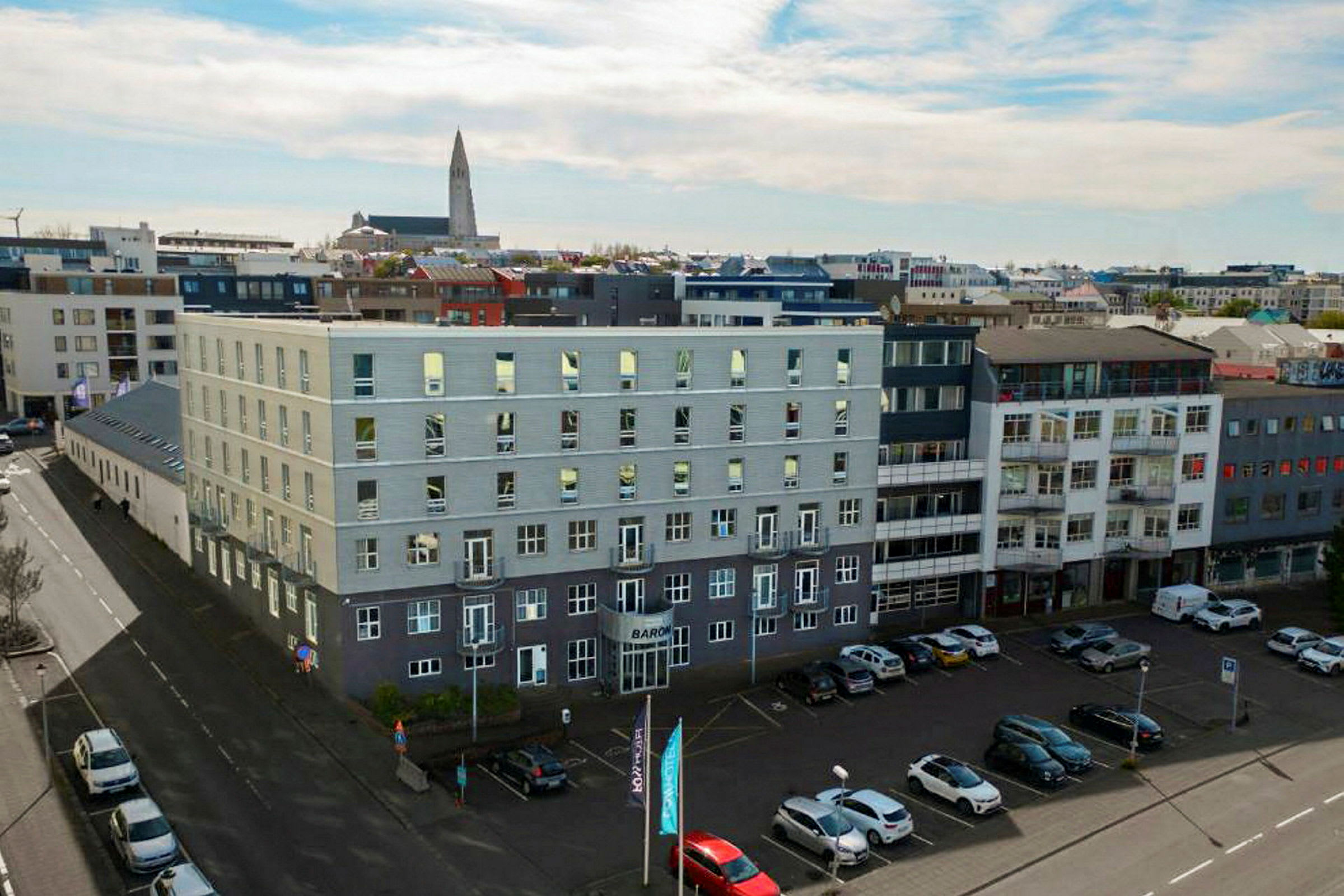 Useat modernit rakennukset ympäröivät avointa pysäköintialueita, jossa on autoja pysäköitynä, ja kirkko näkyy taustalla. (Tekoälyn luoma teksti)