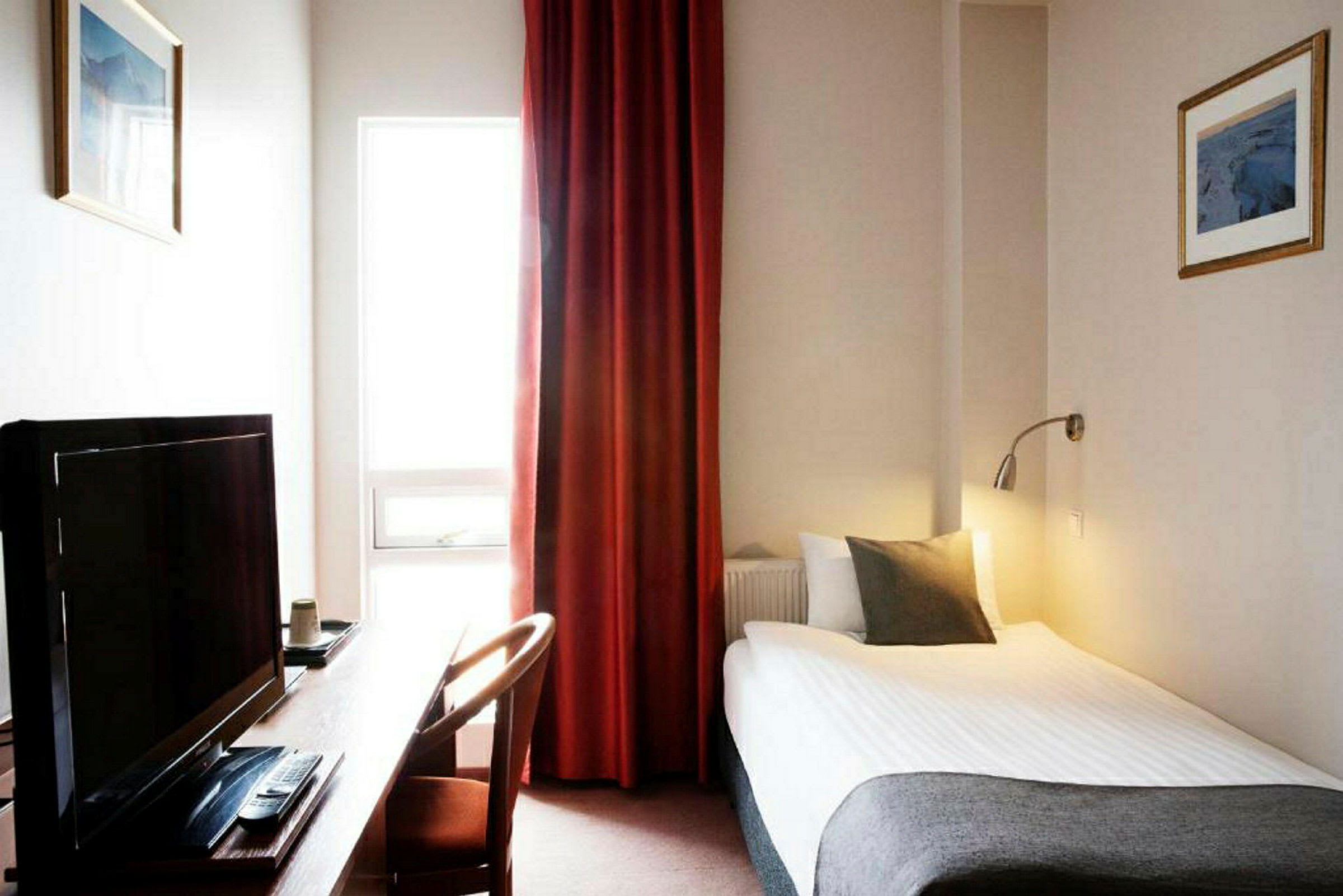 Hotellihuone, jossa on yhden hengen sänky, työpöytä, televisio ja ikkuna punaisilla verhoilla. (Tekoälyn luoma teksti)