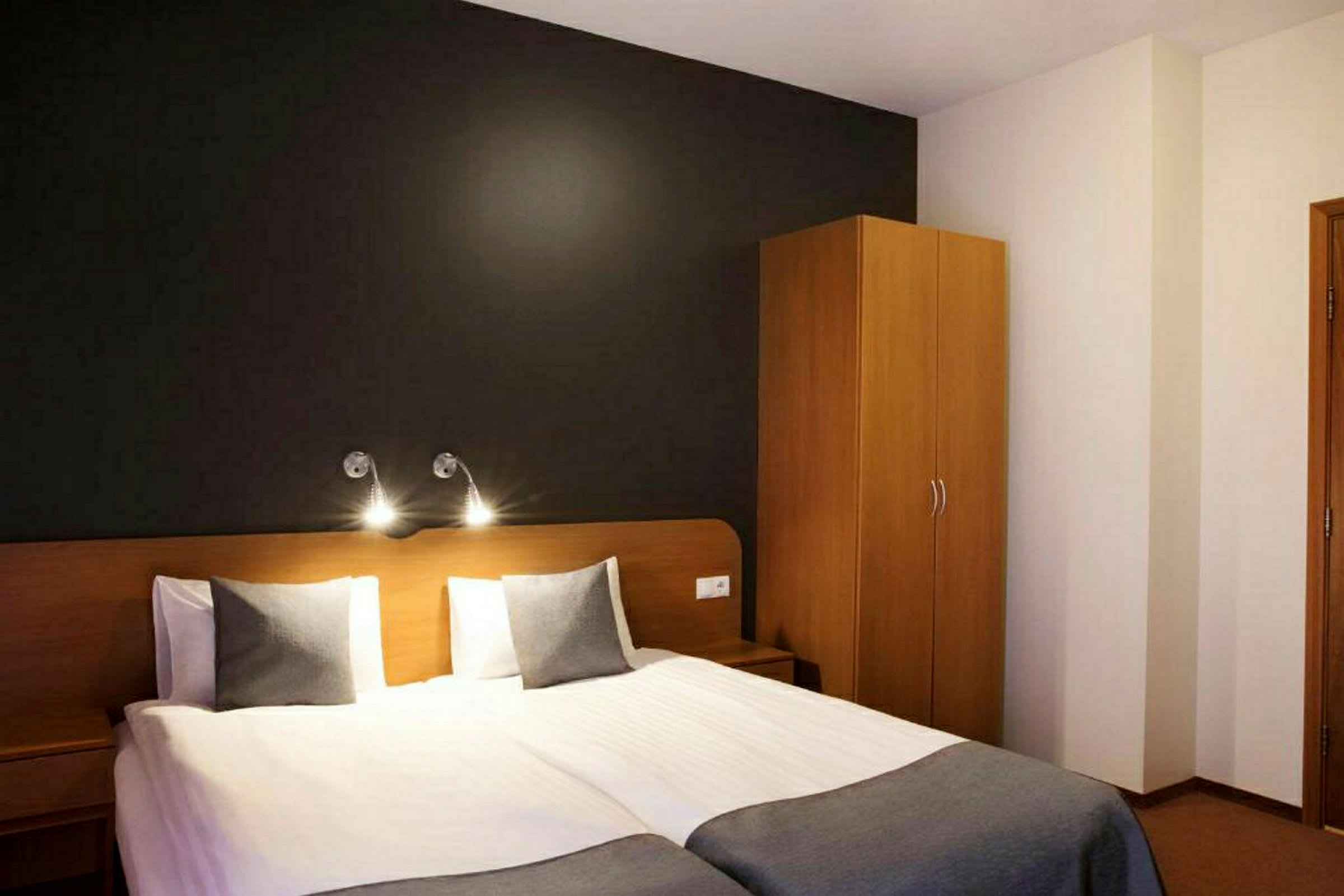 Kuva hotellihuoneesta, jossa on kaksi erillistä sänkyä, tumma seinä takana ja puunvärinen vaatekaappi oikealla. (Tekoälyn luoma teksti)