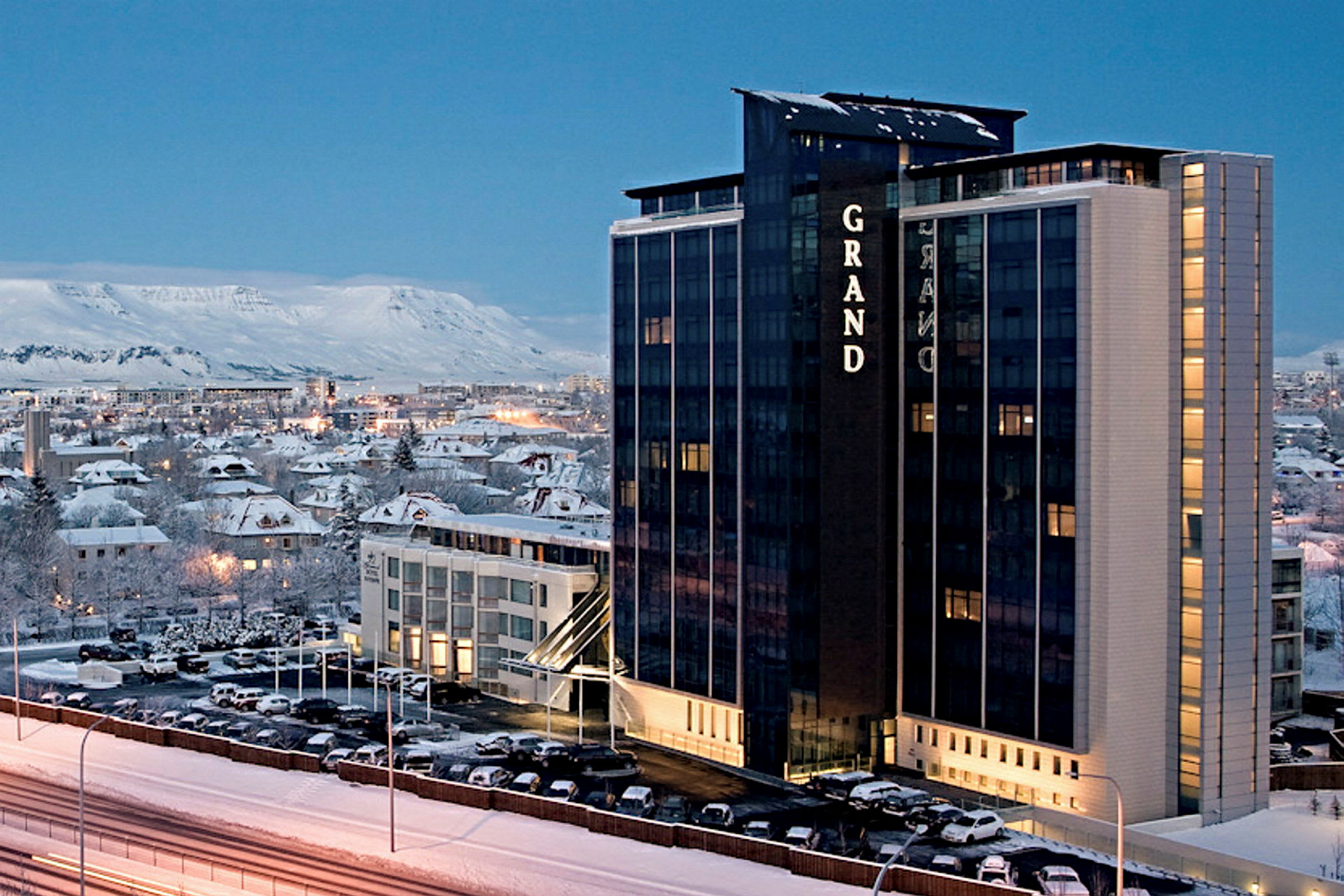 Korkea hotellirakennus talvisessa ympäristössä, taustalla kaupunki ja vuoria. (Tekoälyn luoma teksti)