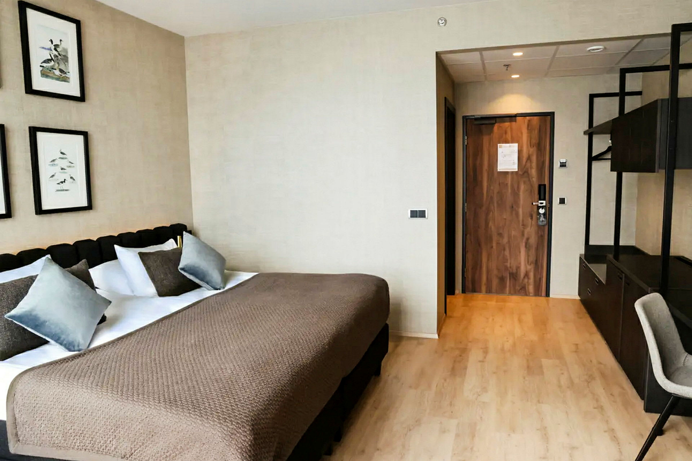 Hotellihuone, jossa on iso sänky, useita tyynyjä ja beige päiväpeite. Yläpuolella seinällä on tauluja, ja oikealla on ovi ulos. (Tekoälyn luoma teksti)