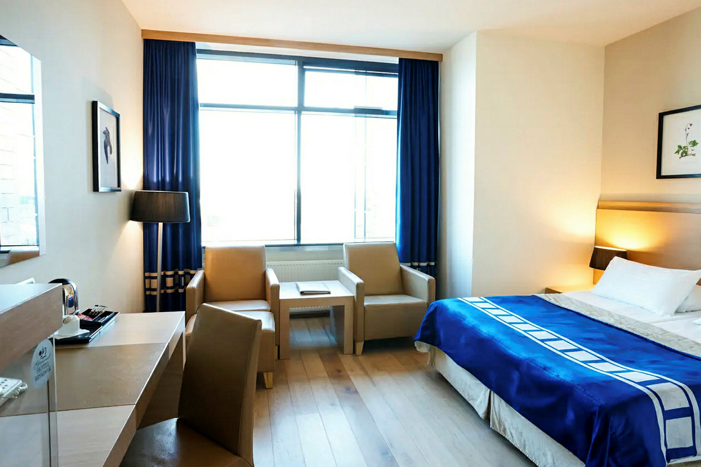 Hotellihuone, jossa on parivuode, sininen päiväpeitto ja kaksi nojatuolia sohvapöydän äärellä. Suuri ikkuna, jossa on siniset verhot, päästää päivänvaloa sisään. (Tekoälyn luoma teksti)