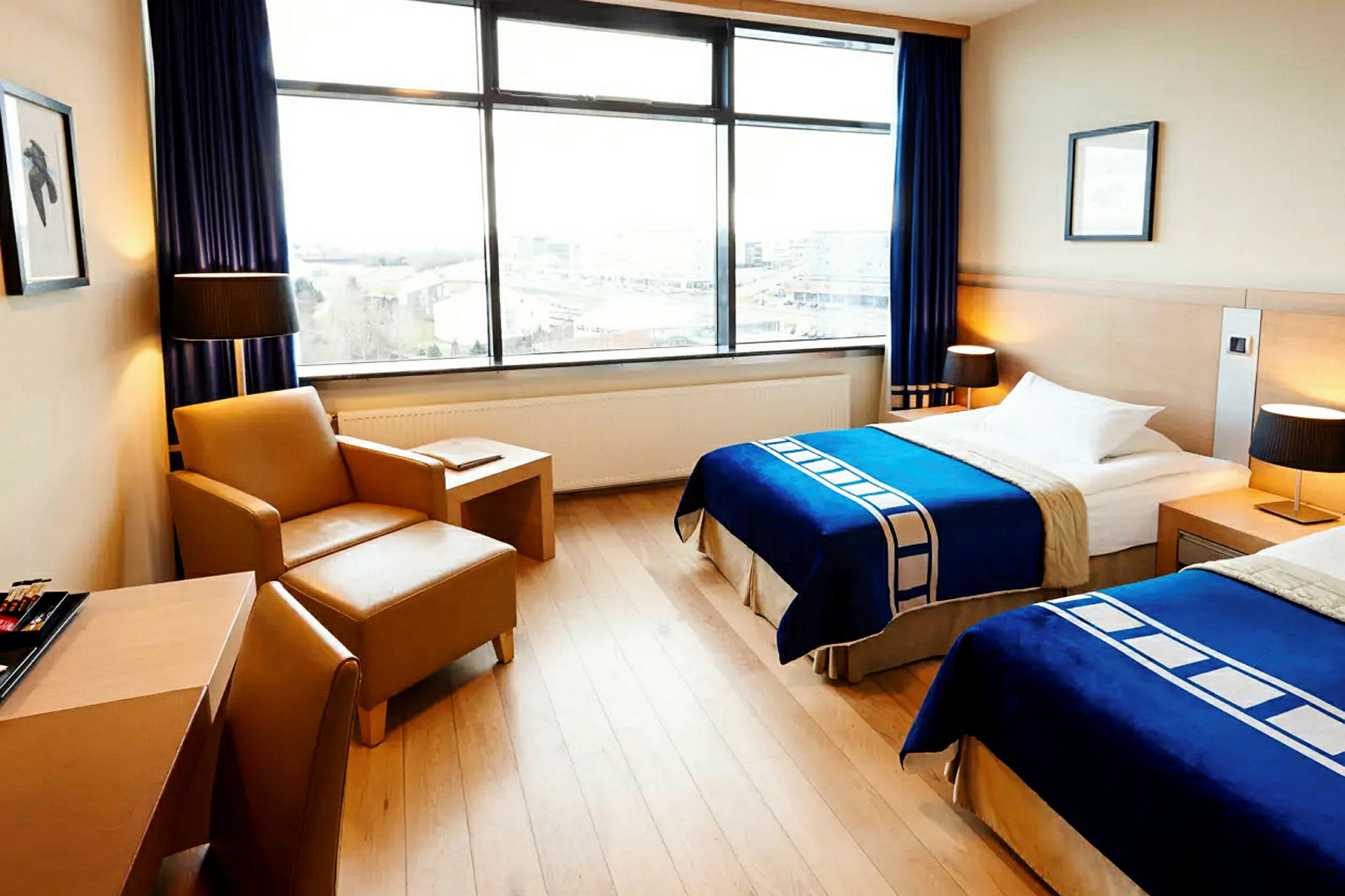Kuva hotellihuoneesta, jossa on kaksi yhden hengen vuodetta, siniset päiväpeitot, nojatuoli pöydän kera ja suuret ikkunat näköalalla. (Tekoälyn luoma teksti)