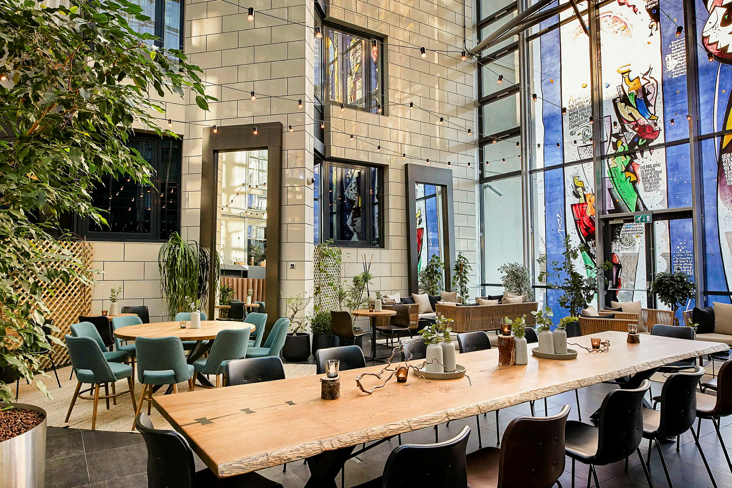 Sisälounge, jossa on suuret ikkunat, istuinalueita useiden pöytien ympärillä ja vihreitä kasveja eri kohdissa. (Tekoälyn luoma teksti)