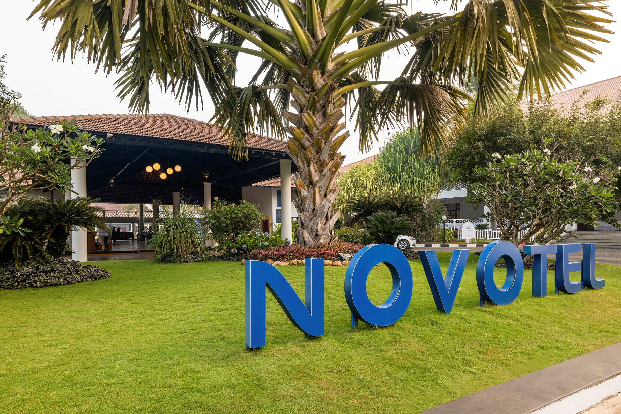 Puutarha, jossa on suuri palmu ja vihreä nurmikko rakennuksen edessä, sininen kyltti jossa lukee "Novotel". (Tekoälyn luoma teksti)