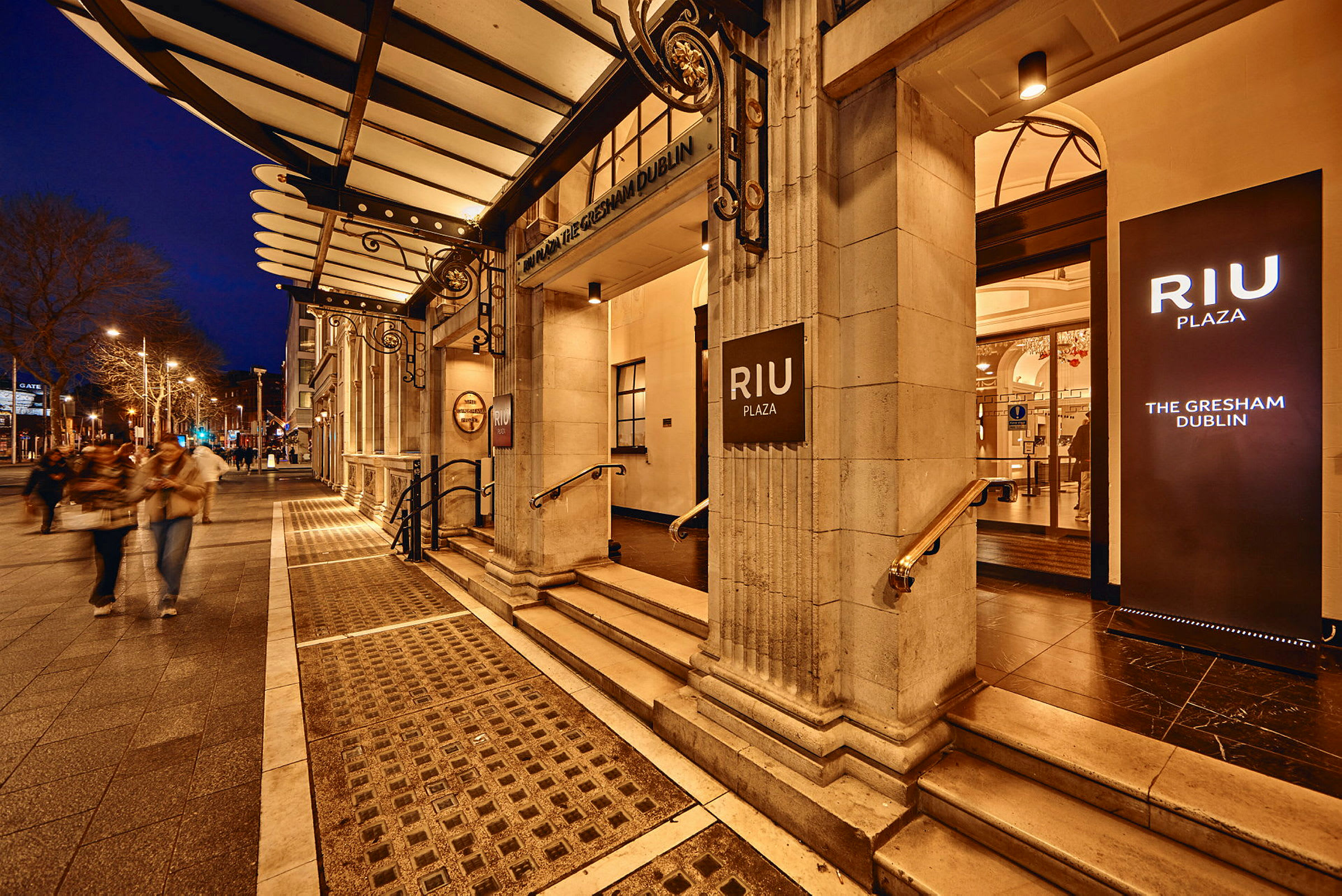 Hotel RIU Plaza Gresham Dublin sisäänkäynti valaistuilla kylteillä, iltamaisema. (Tekoälyn luoma teksti)