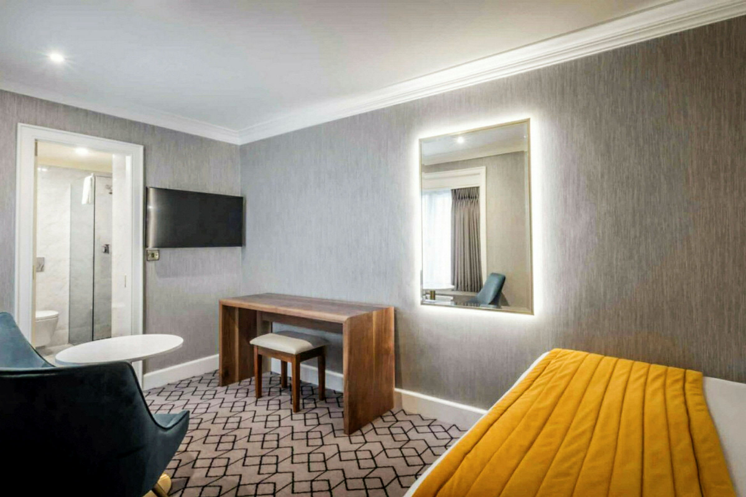 Hotellihuone, jossa harmaat seinät, peili, kirjoituspöytä, tuoli ja vuode, jossa keltainen viltti. (Tekoälyn luoma teksti)