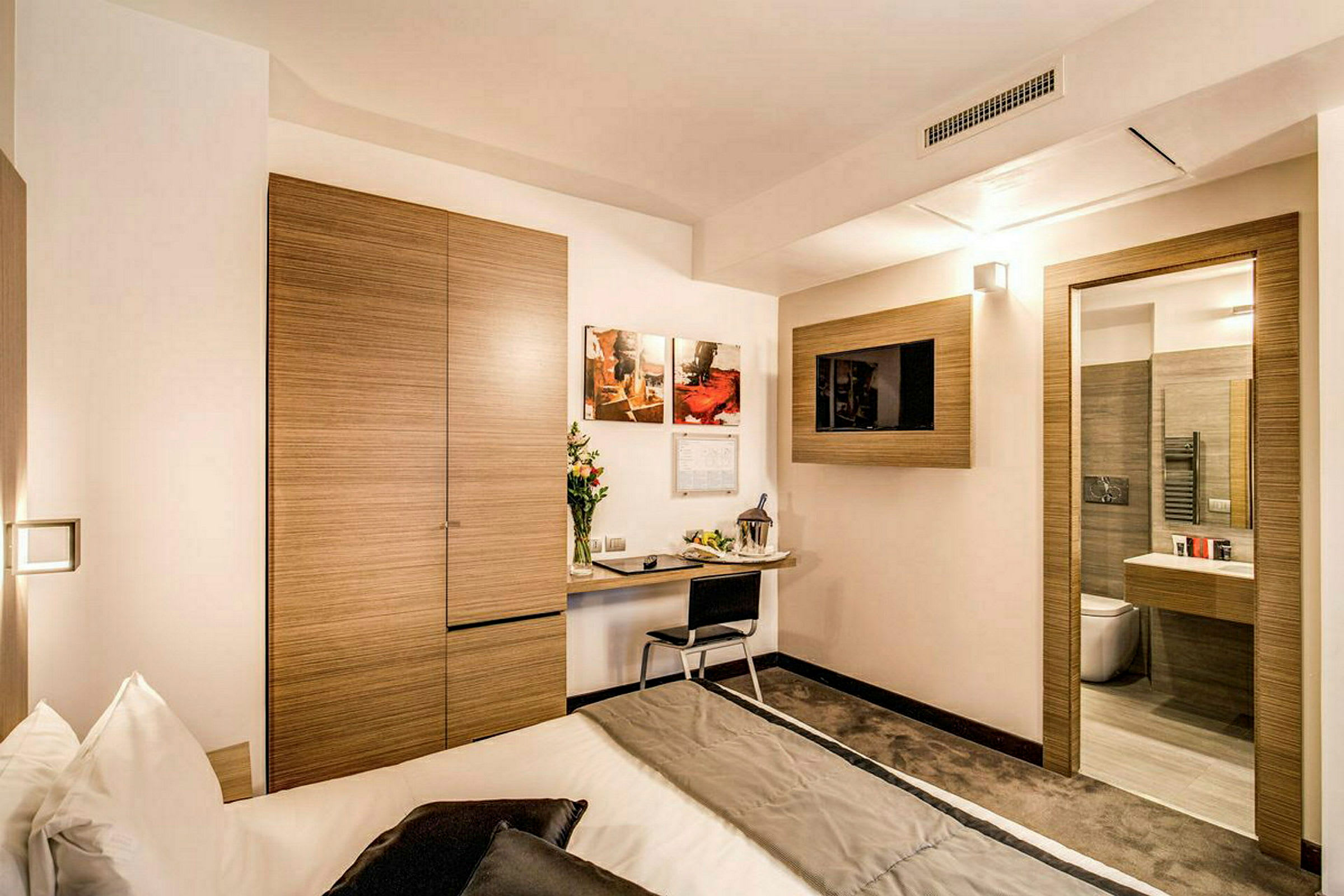 Moderni hotellihuone, jossa on kirjoituspöytä, sänky ja seinälle asennettu televisio. (Tekoälyn luoma teksti)