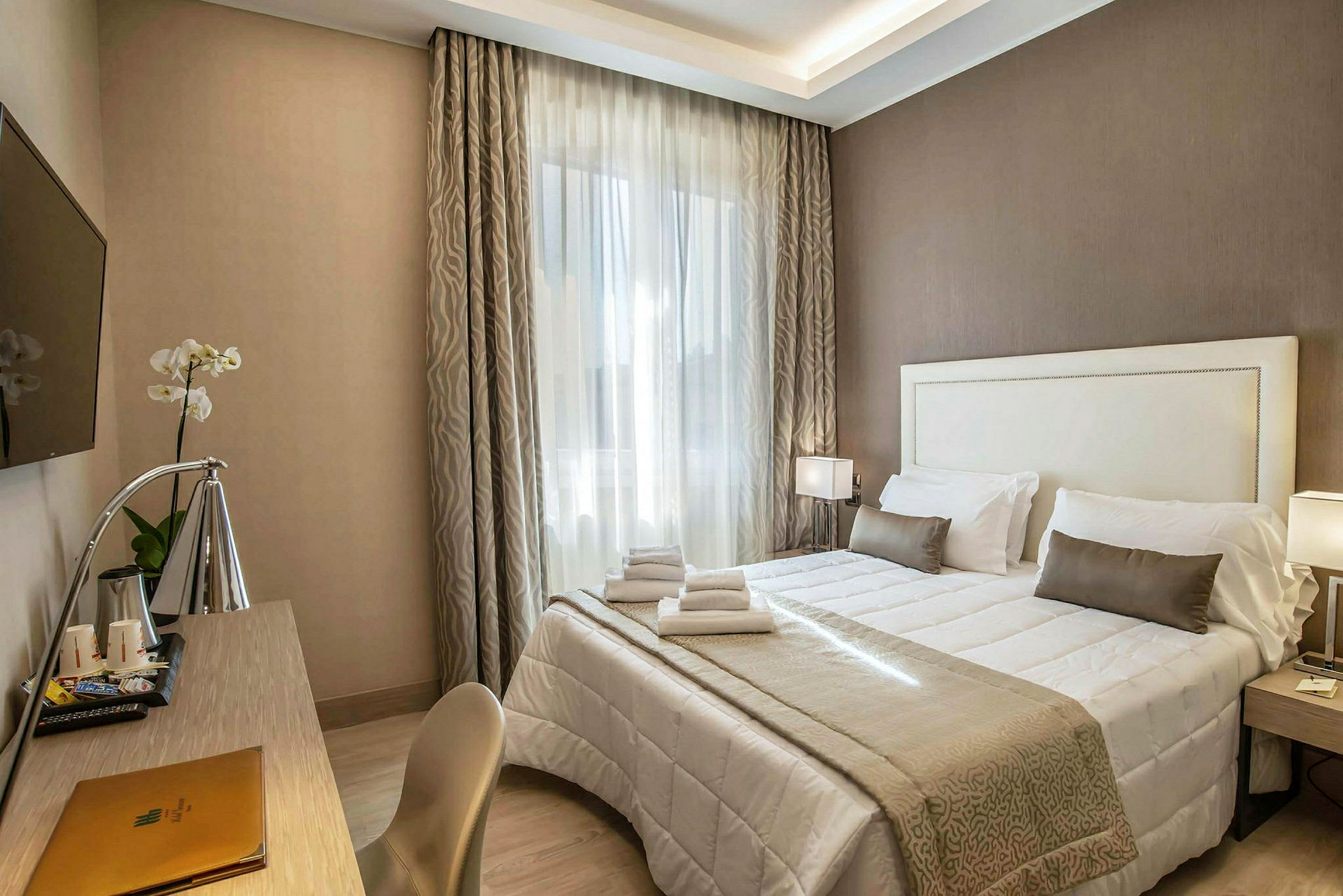 Hotellihuone, jossa on parivuode, valkoiset ja beige lakanat, kirjoituspöytä, lamppu ja seinälle kiinnitettävä TV. (Tekoälyn luoma teksti)