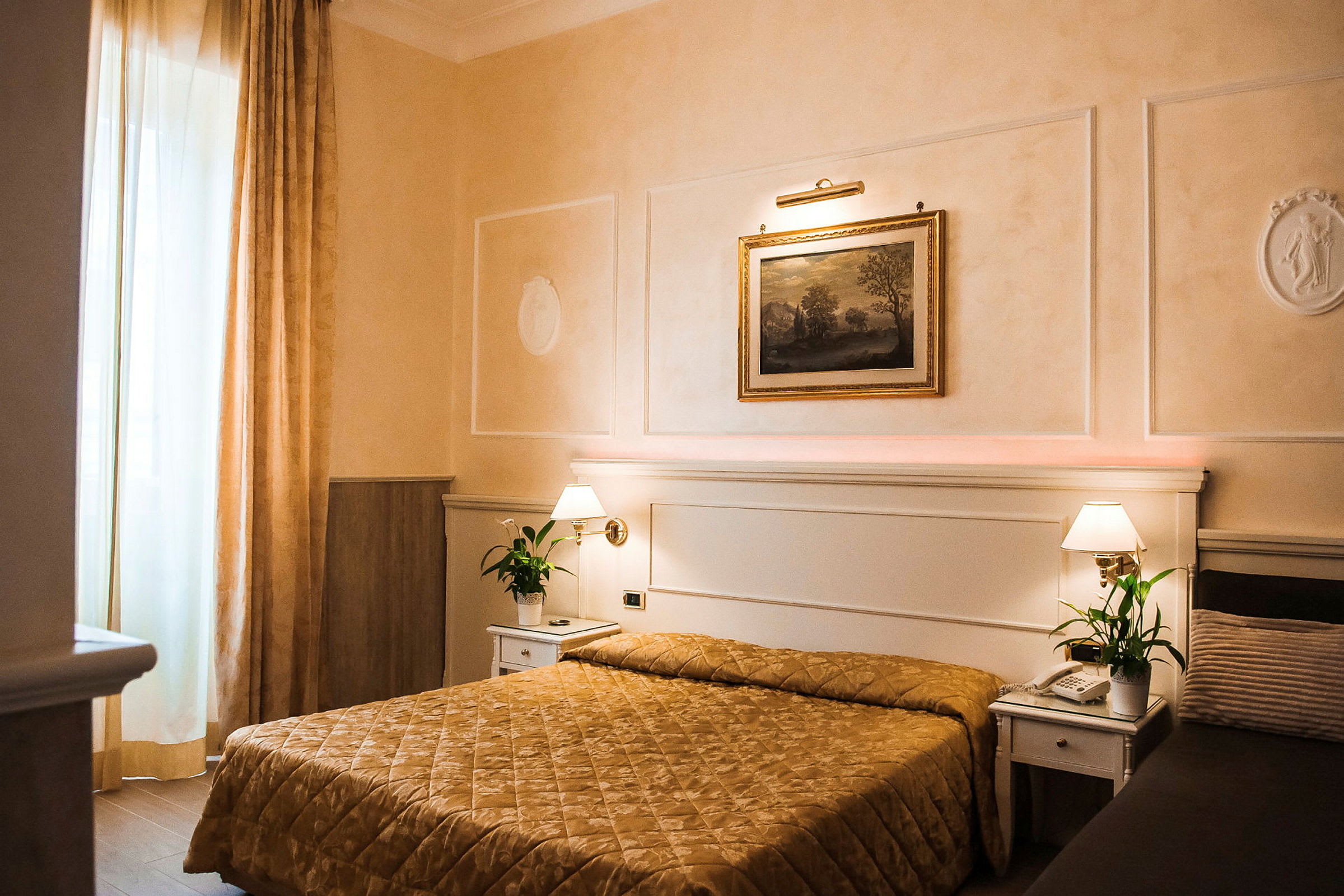 Hotellihuone, jossa on sänky kultaisen värisellä peitteellä, kaksi yöpöytää lampuilla ja vihreillä kasveilla sekä seinällä sängyn yläpuolella oleva taulu. (Tekoälyn luoma teksti)