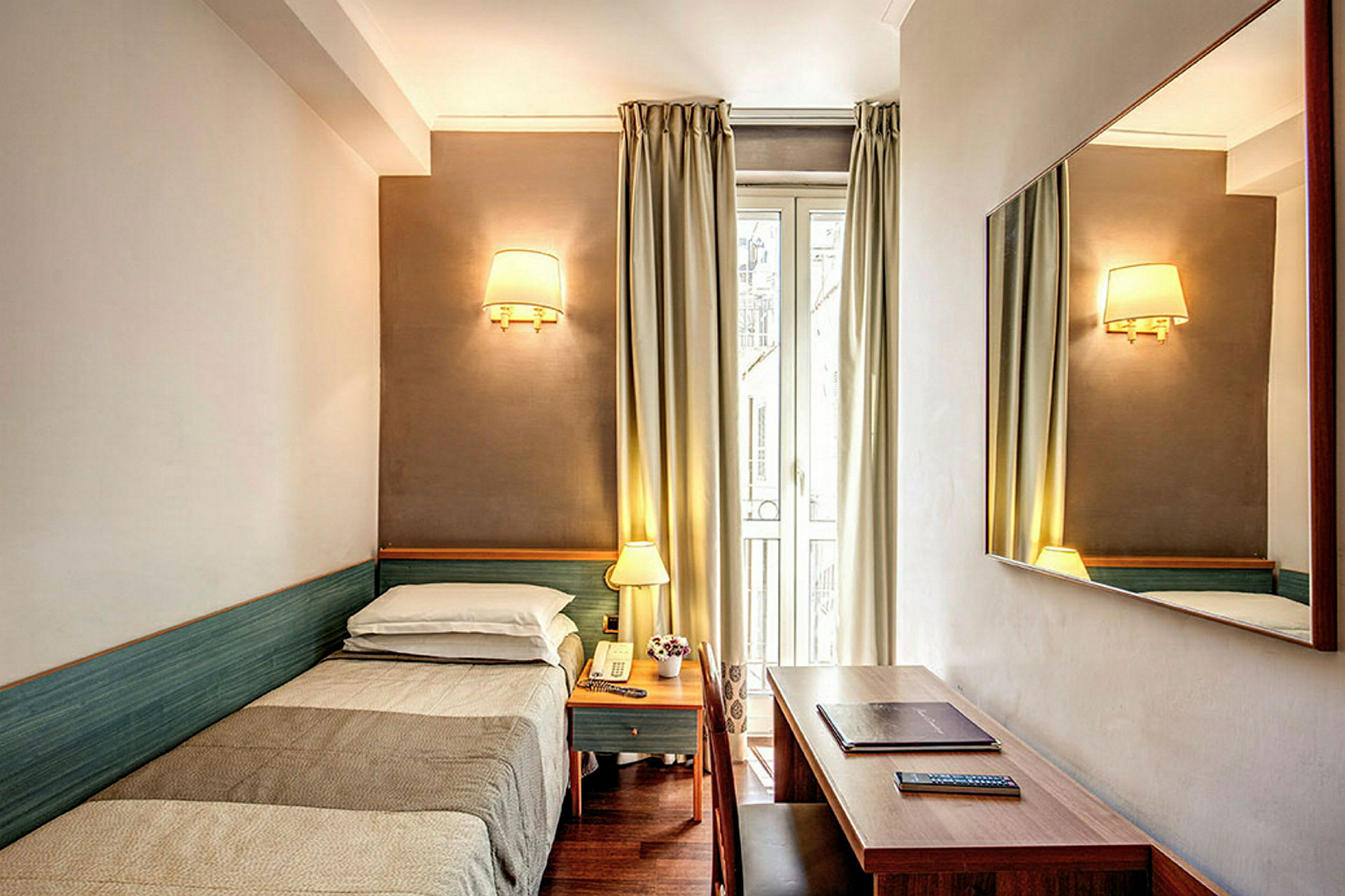 Hotellihuone, jossa on yhden hengen sänky, yöpöytä, seinällä oleva peili ja kirjoituspöytä. (Tekoälyn luoma teksti)