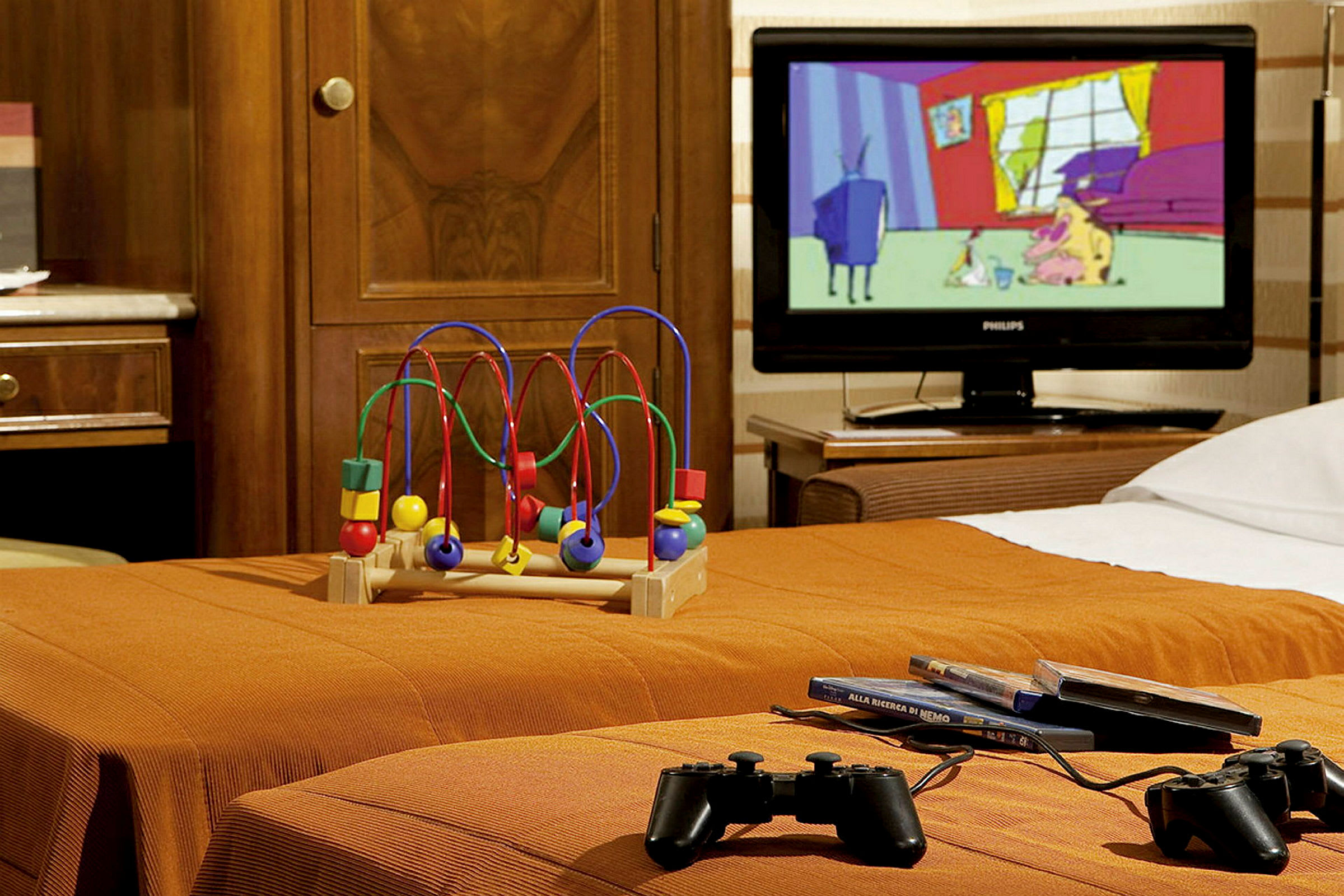 Hotellihuone, jossa on lelu, televisio ja peliohjaimet oranssilla sängyllä. (Tekoälyn luoma teksti)
