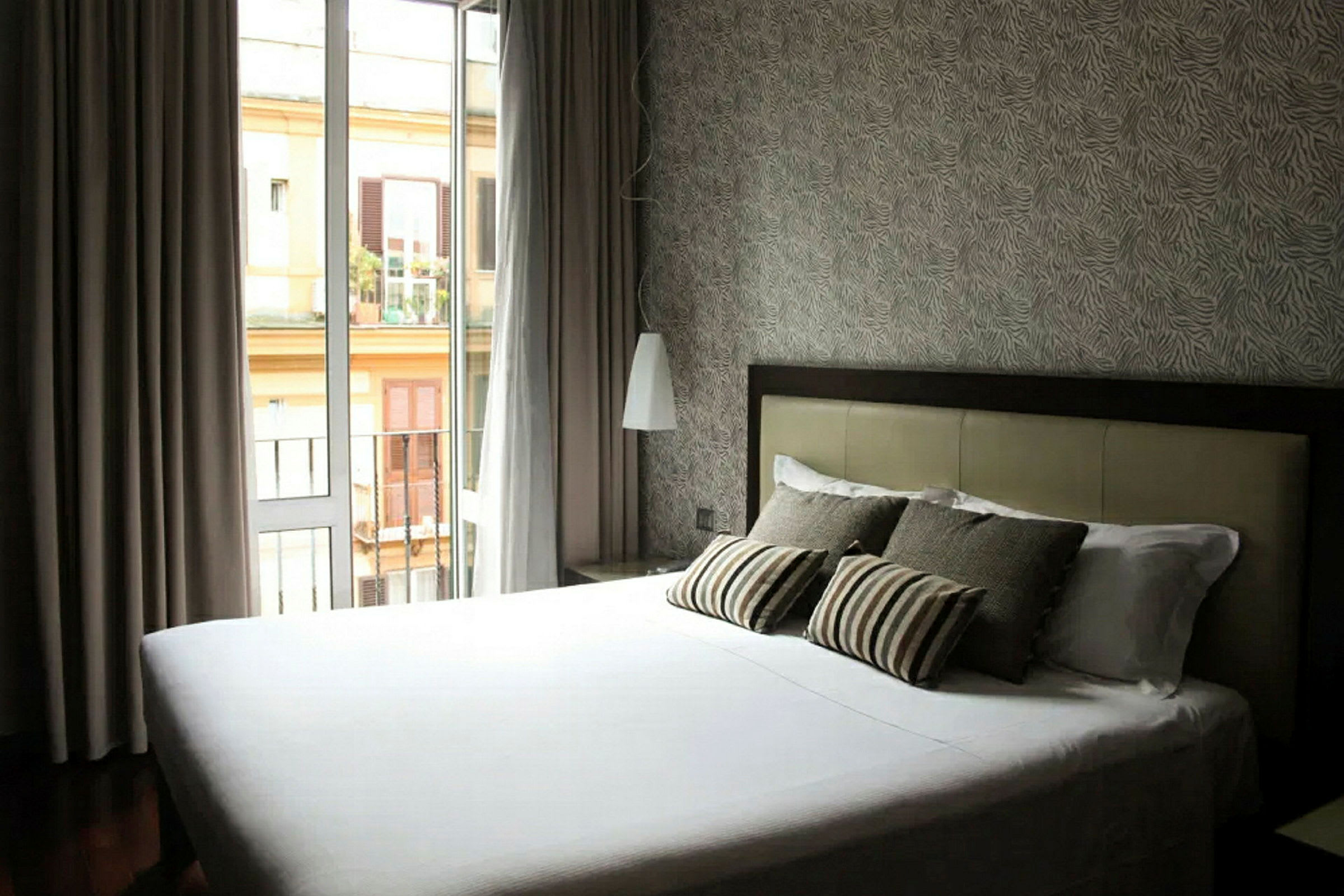 Hotellihuone, jossa on valkoisilla lakanoilla varustettu sänky ja useita tyynyjä, läheinen parvekkeen ovi päästää luonnonvaloa sisään ja seinällä on yksityiskohtainen tapetti. (Tekoälyn luoma teksti)