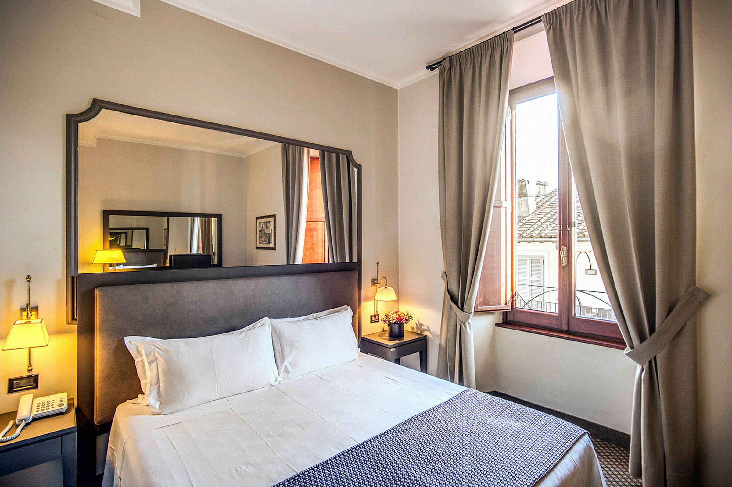 Hotellihuone, jossa parivuode, yöpöytä, peili ja ikkuna beigeillä verhoilla. (Tekoälyn luoma teksti)