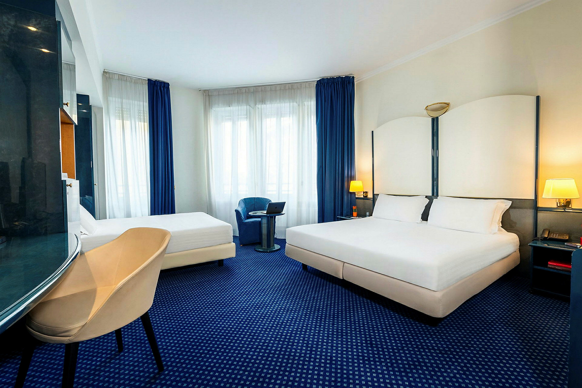 Hotellihuone, jossa on parivuode ja yhden hengen vuode, nojatuoli ja ikkuna sinisillä verhoilla. (Tekoälyn luoma teksti)