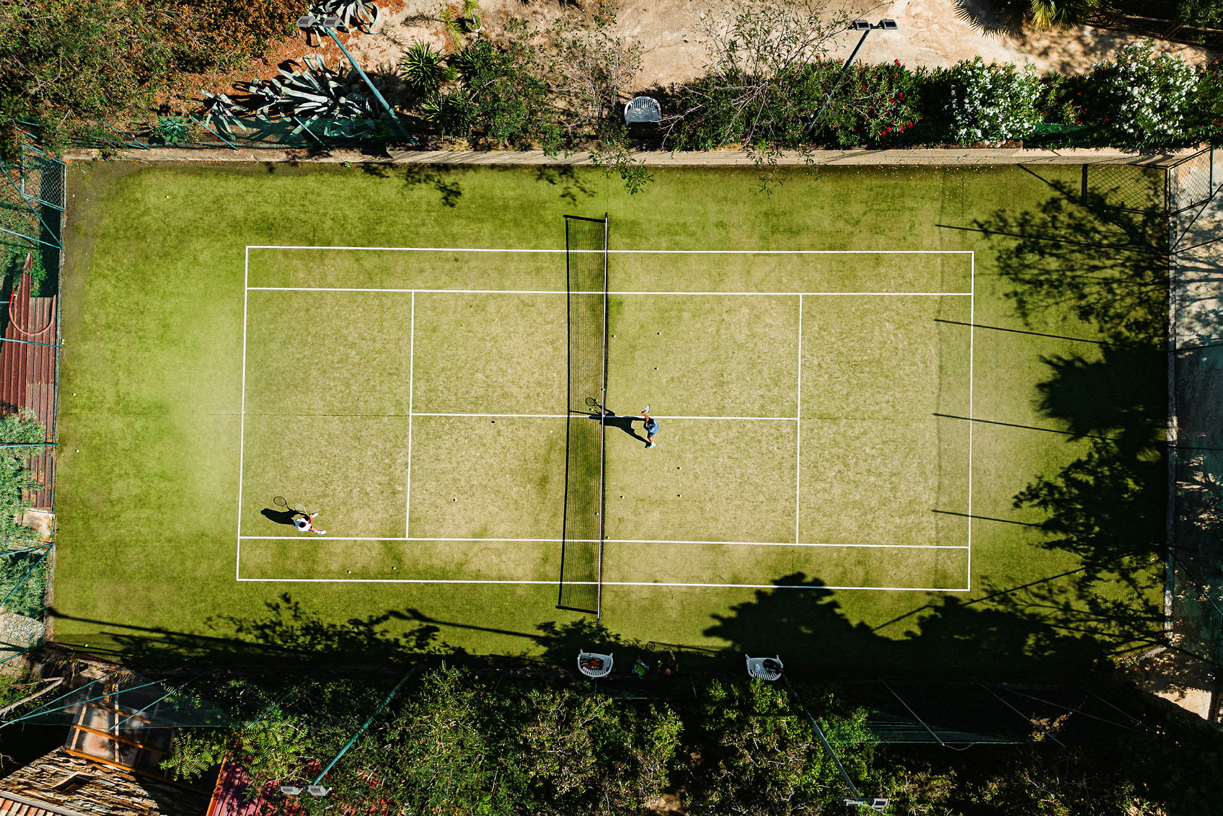 Ilmakuva kahdesta henkilöstä pelaamassa tennistä hiekkaisella kentällä kasvillisuuden ympäröimänä. (Tekoälyn luoma teksti)