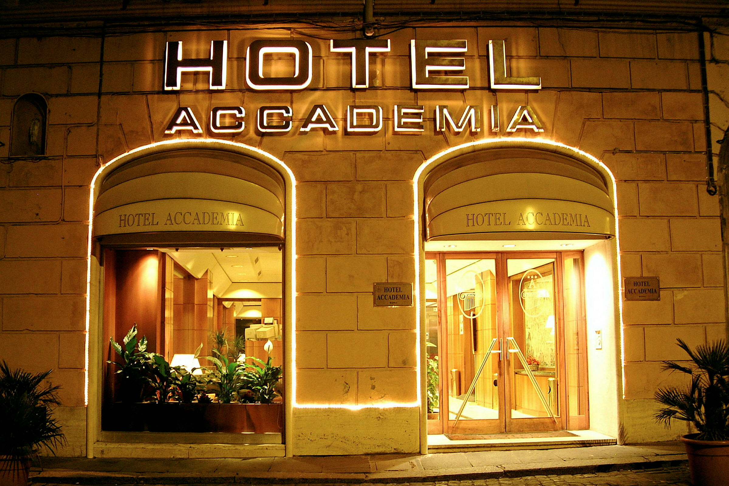 Accademia-hotellin valaistu sisäänkäynti, lasiovet ja kasvit. (Tekoälyn luoma teksti)