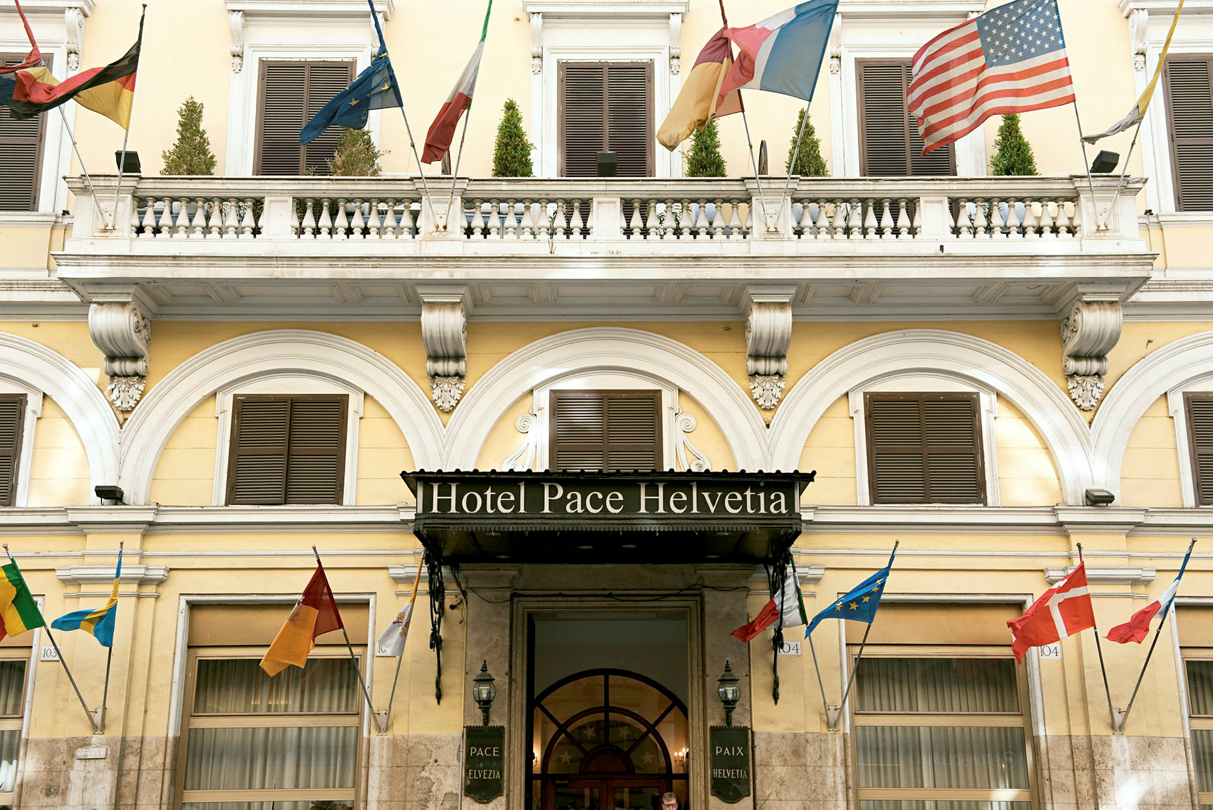 Hotellin julkisivu, jossa nimi 'Hotel Pace Helvetia', koristeltu monien maiden lipuilla. (Tekoälyn luoma teksti)