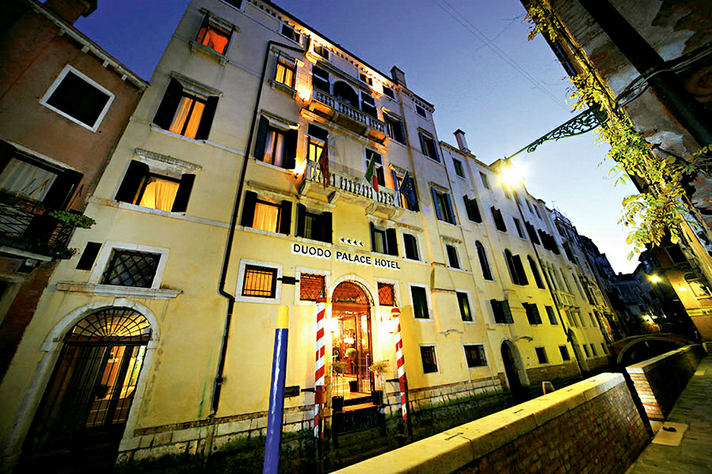 Kolmikerroksinen asuinrakennus, jossa kyltti Duodo Palace Hotel, kanavan ja kävelytien ympäröimänä. (Tekoälyn luoma teksti)