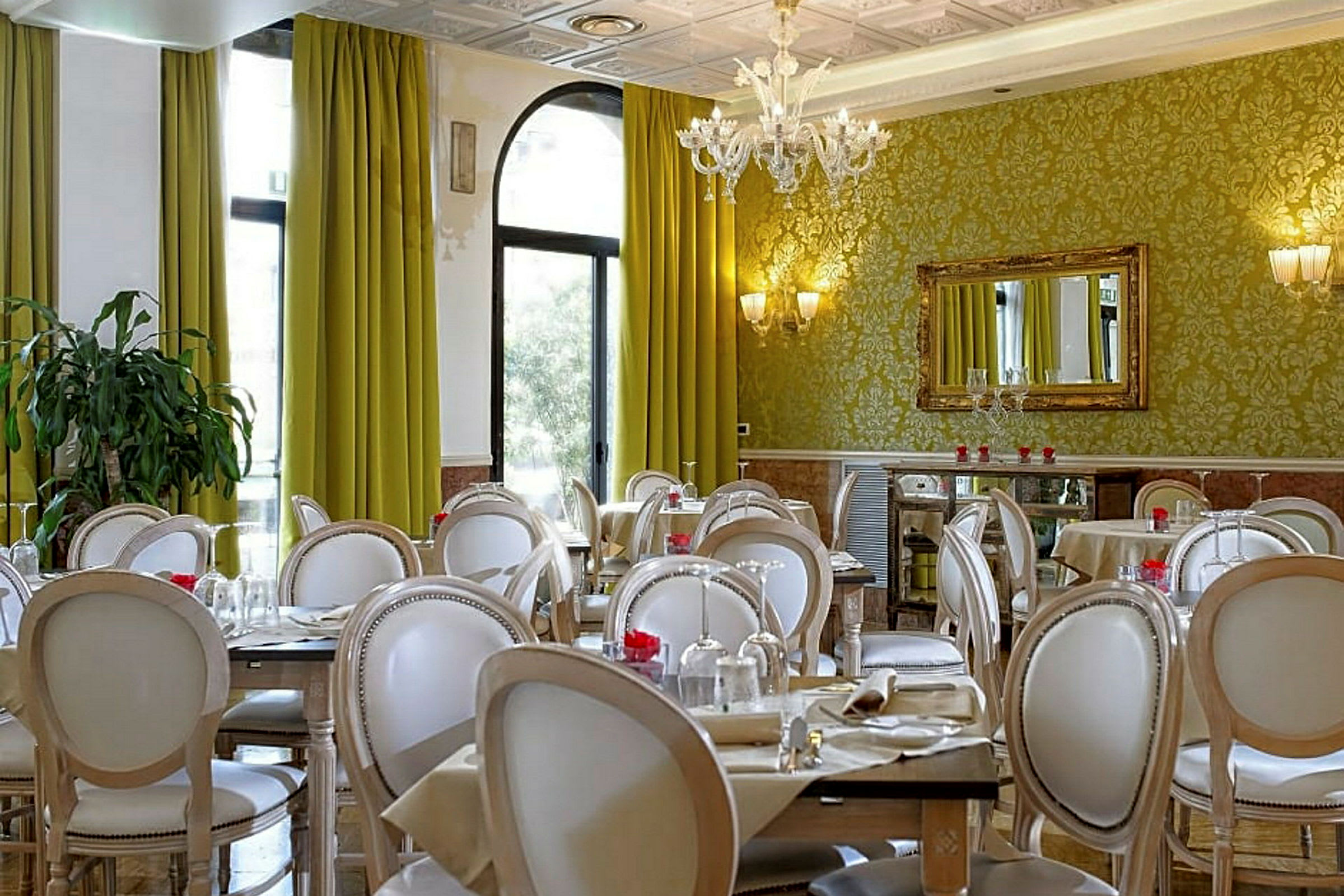 Elegantly sisustettu ravintola-alue vihreillä verhoilla, koristeellisilla seinillä ja valkoisilla tuoleilla katettujen pöytien ympärillä. (Tekoälyn luoma teksti)