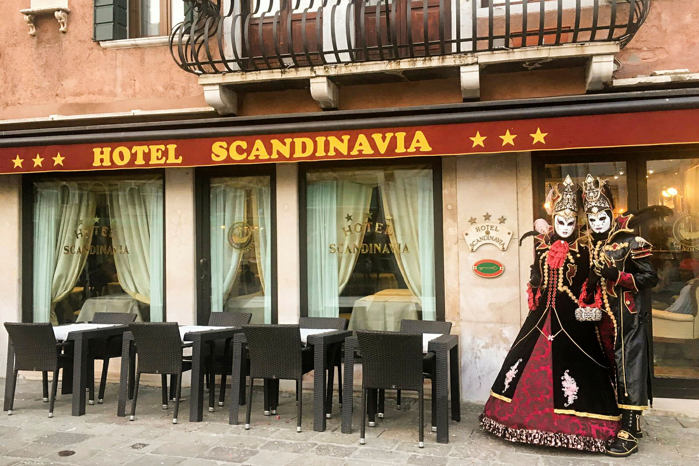 Hotellin julkisivu, jossa lukee 'Hotel Scandinavia', ulkona kalustetut istumapaikat ja perinteisissä naamiaisasuissa olevia henkilöitä. (Tekoälyn luoma teksti)