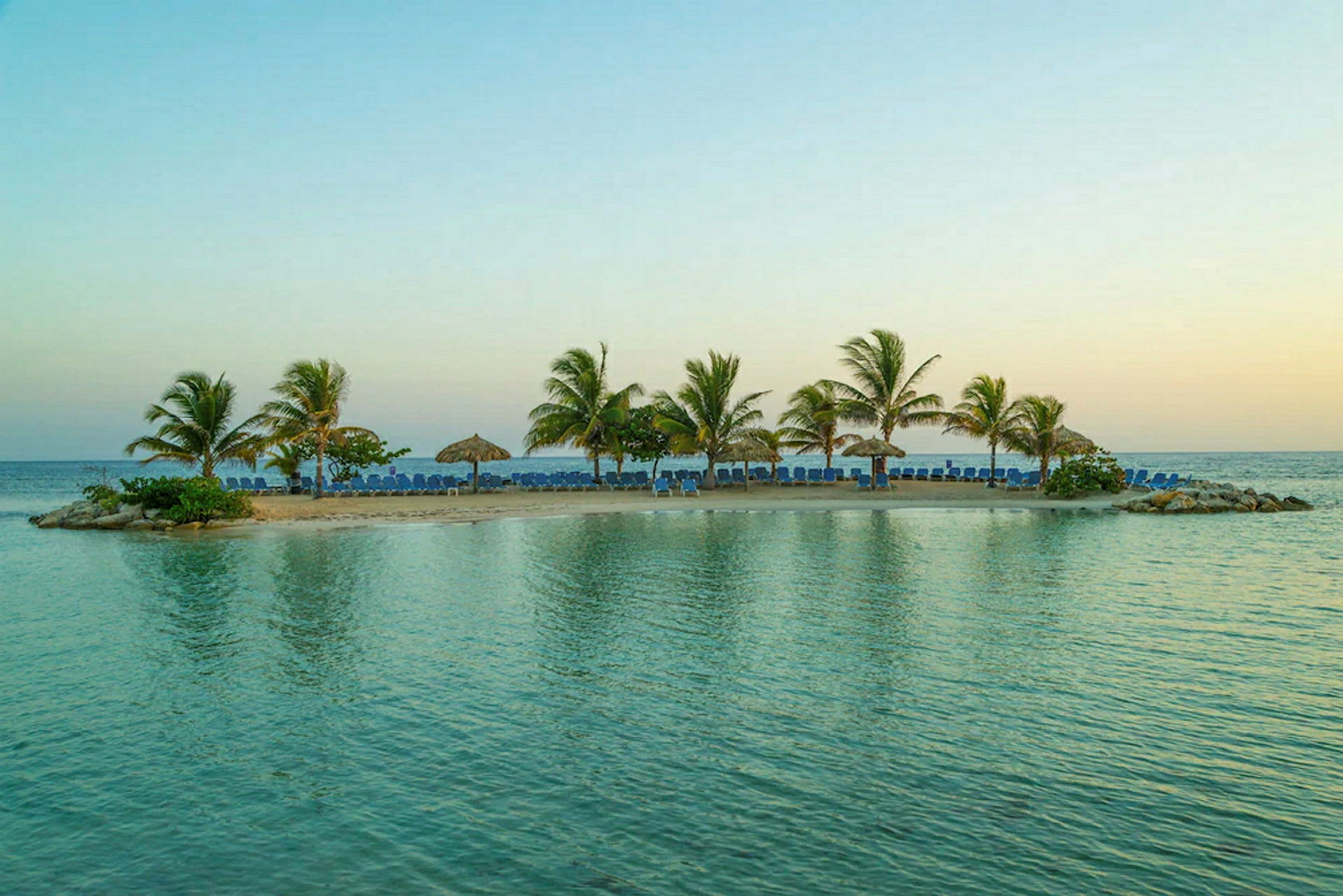 Pieni saari, jolla on hiekkaranta, palmuja ja aurinkotuoleja, sinisen veden ympäröimänä. (Tekoälyn luoma teksti)