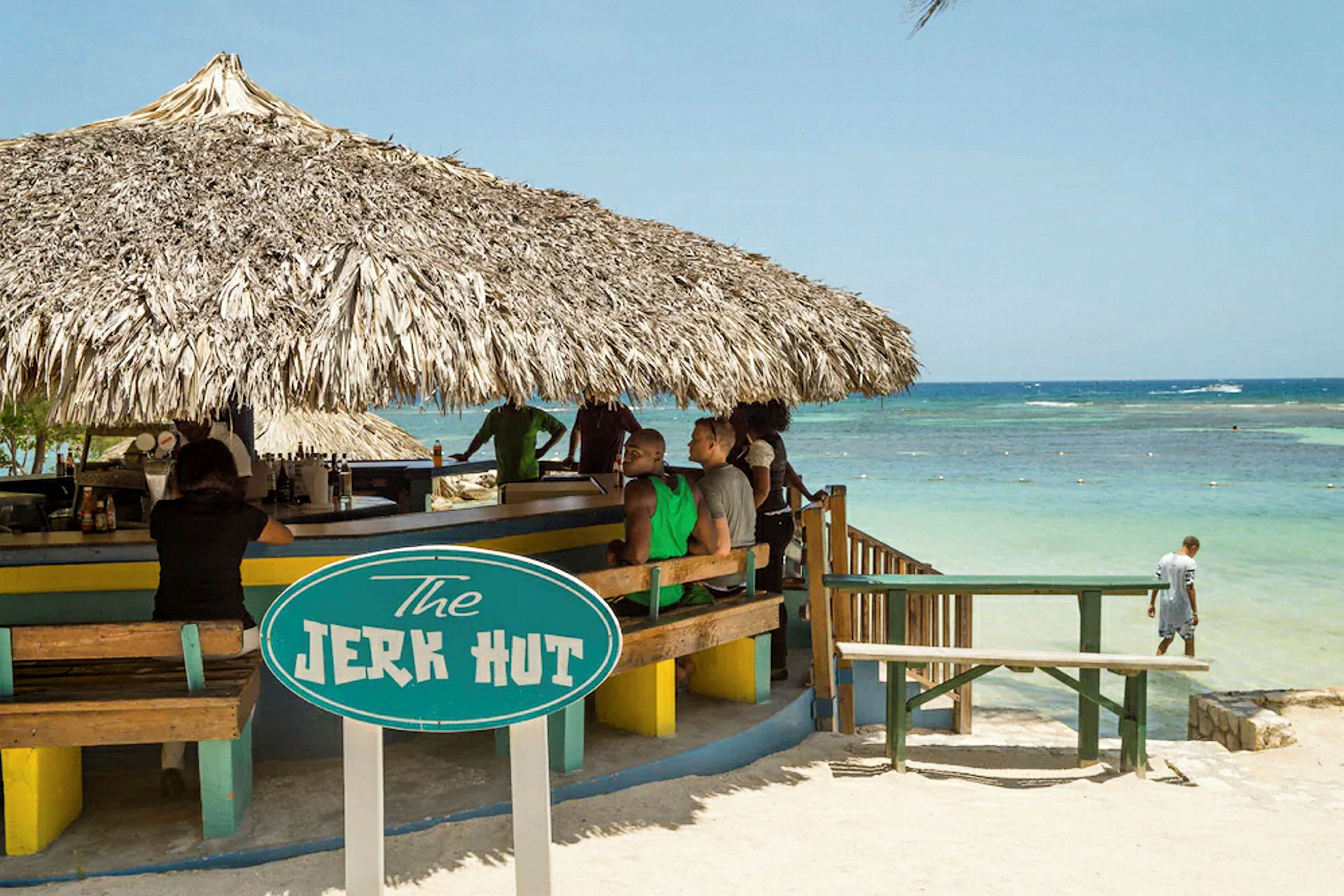 Rantabaari, jossa on olkikatto ja kyltti, jossa lukee "The Jerk Hut". Taustalla näkyy meri. (Tekoälyn luoma teksti)