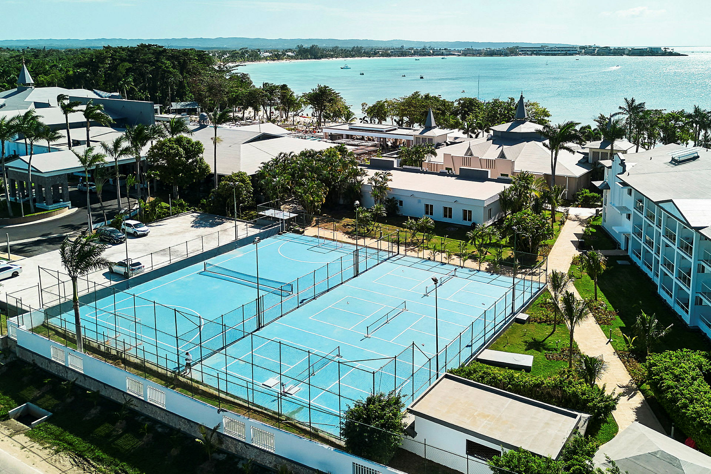 Näköala sinisen pinnan tenniskentälle, jota ympäröivät rakennukset ja vihreät alueet lähellä merta. (Tekoälyn luoma teksti)