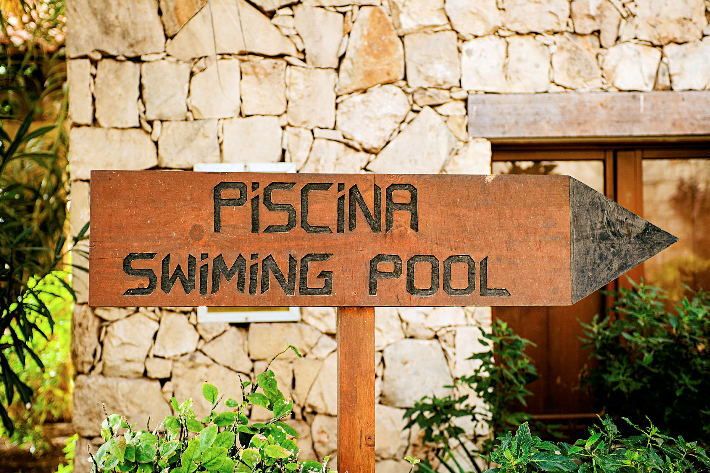 Kyltti, jossa lukee 'Piscina - Swimming Pool', kivimuurin edessä. (Tekoälyn luoma teksti)