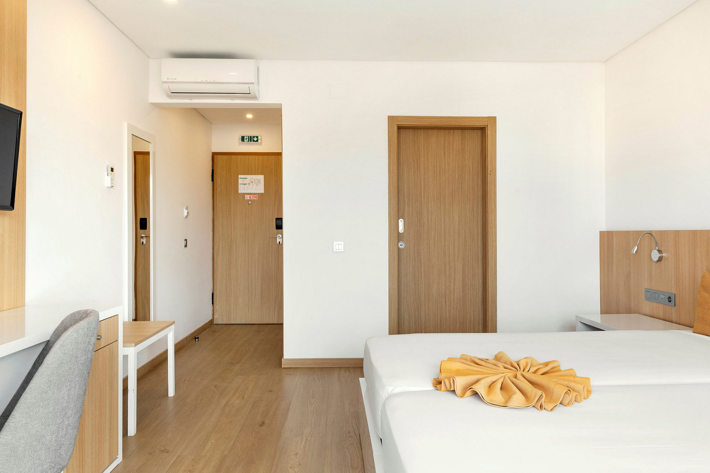 Hotellihuone, jossa on pedattu sänky ja kirjoituspöytä harmaan tuolin vieressä, puuovet ja seinälle asennettu ilmastointilaite. (Tekoälyn luoma teksti)