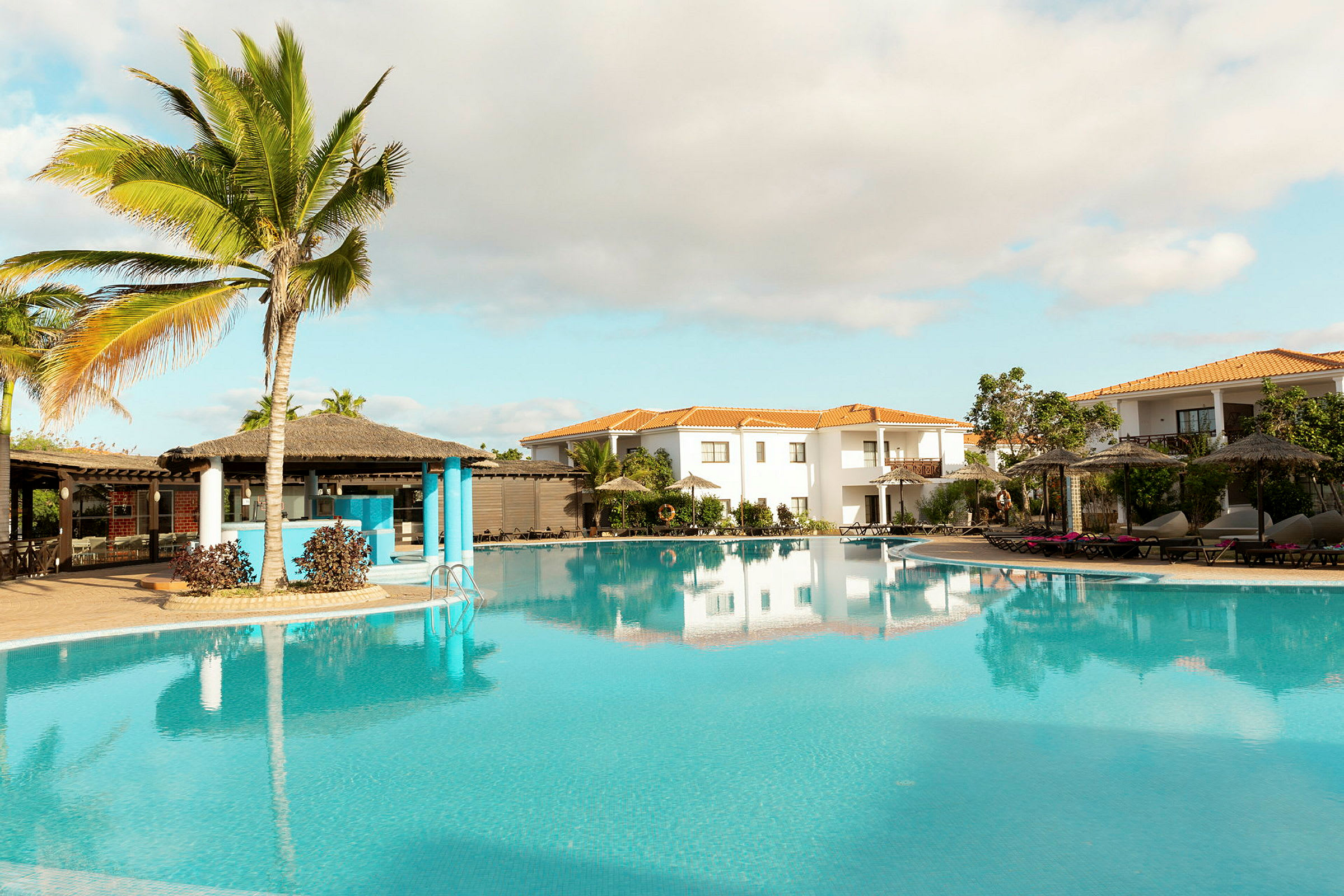 Trooppinen uima-allasalue palmujen, varjojen ja ympäröivien rakennusten kanssa. (Tekoälyn luoma teksti)