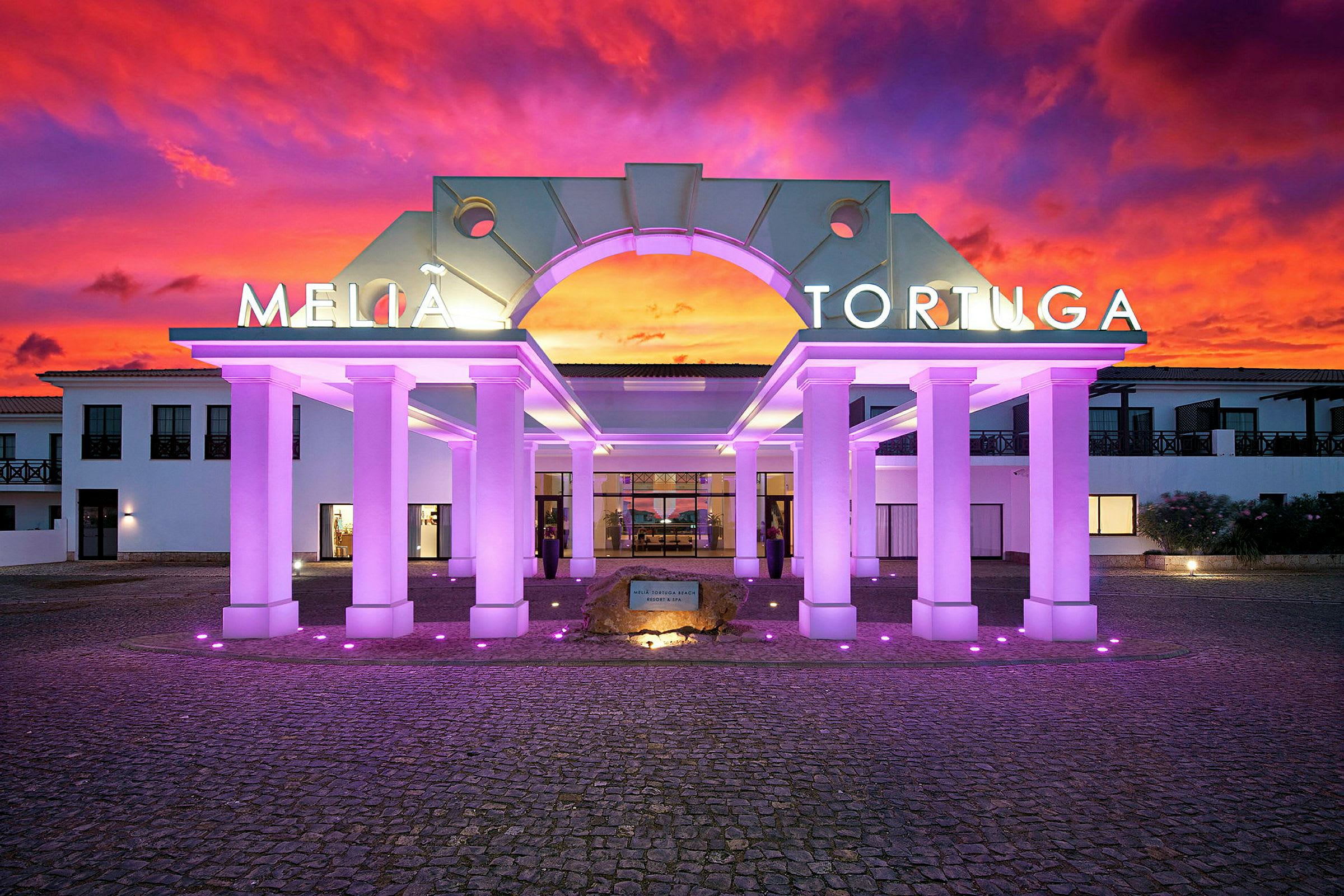 Hotellin sisäänkäynti valaisimilla pylväillä ja värikäs auringonlasku taustalla. (Tekoälyn luoma teksti)