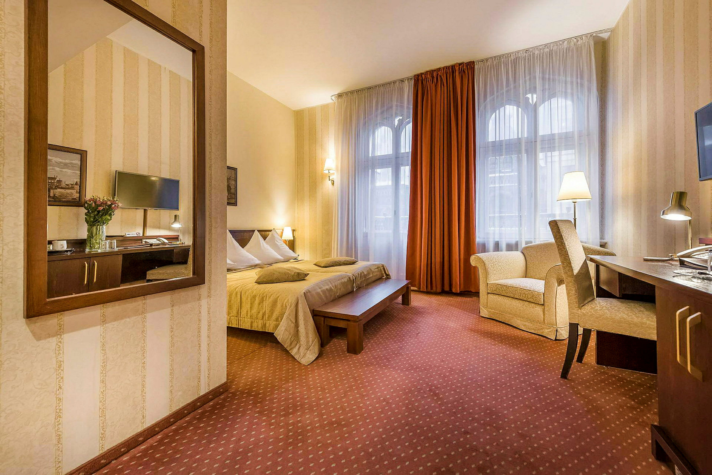 Hotellihuone, jossa on parivuode, kirjoituspöytä ja tuoli sekä nojatuoli. Ikkunat verhoilla ja seinäpeili. (Tekoälyn luoma teksti)