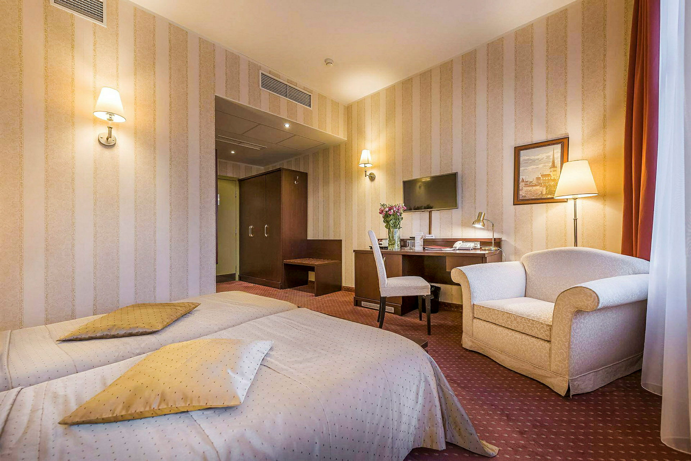Hotellihuone, jossa kaksi erillistä vuodetta, kirjoituspöytä tuoleineen, seinälle kiinnitetty televisio ja nojatuoli. (Tekoälyn luoma teksti)